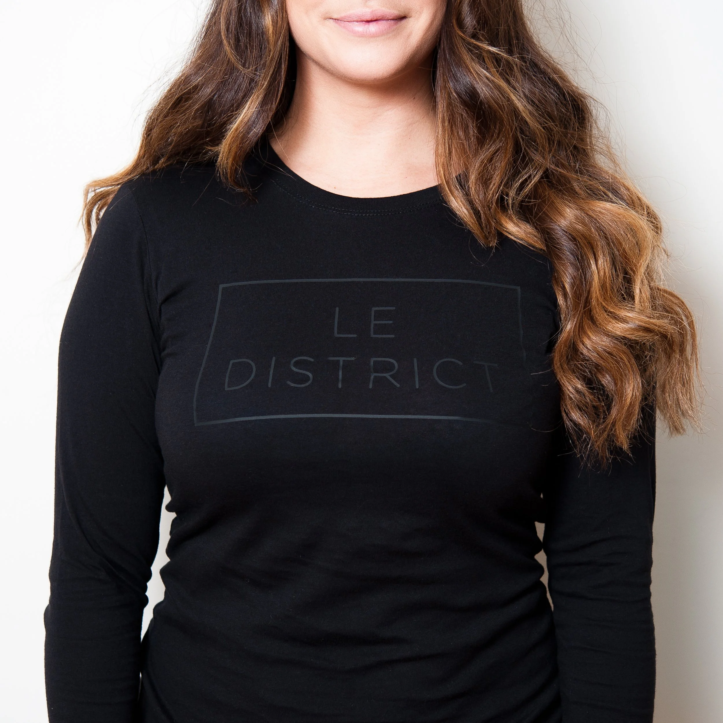 Close-up of a woman's face and upper body, wearing a black shirt with a graphic design, and long wavy brown hair, against a plain light background.