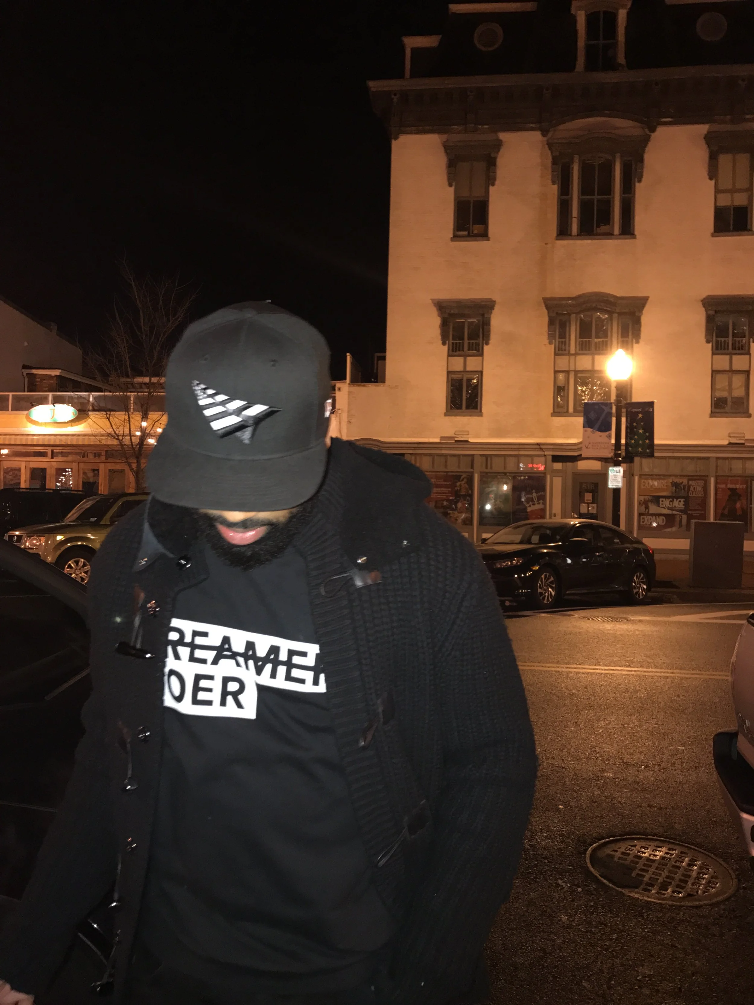 A man wearing a dark baseball cap and black hoodie with white text on it standing outdoors at night in front of a building with lit windows and parked cars.