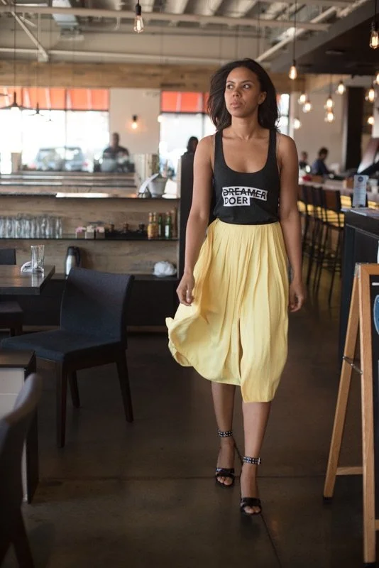 A woman walking inside a modern restaurant or café with wooden accents and ambient lighting. She is wearing a black tank top with the words 'DREAMER DOER' and a flowing yellow skirt, paired with black high-heeled sandals.