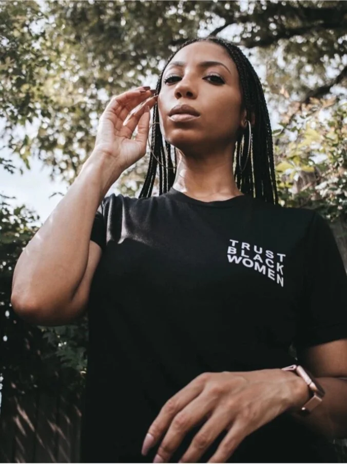 A woman with braided hair and hoop earrings standing outdoors, wearing a black T-shirt with the text 'TRUST BLACK WOMEN'.