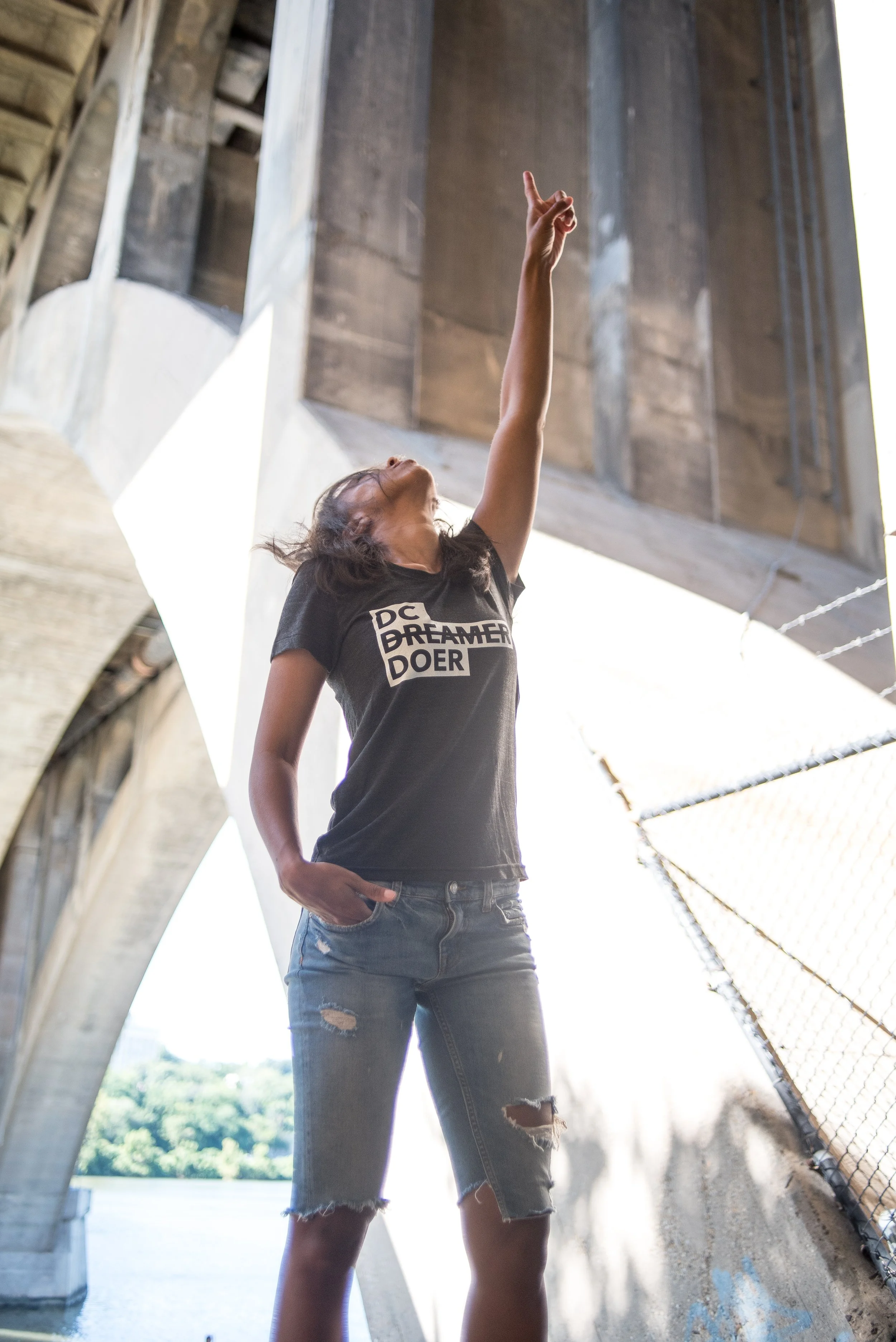 A young woman standing underneath a bridge with an industrial concrete structure, pointing upward with her right hand, wearing a black T-shirt that reads 'DC Dreamer Doer' and ripped jeans.