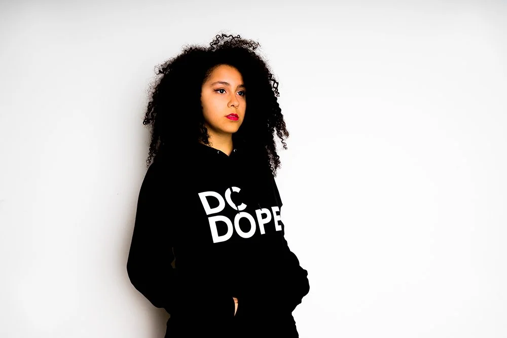 Young woman with curly black hair and red lipstick wearing a black hoodie with white text, standing against a plain light background.