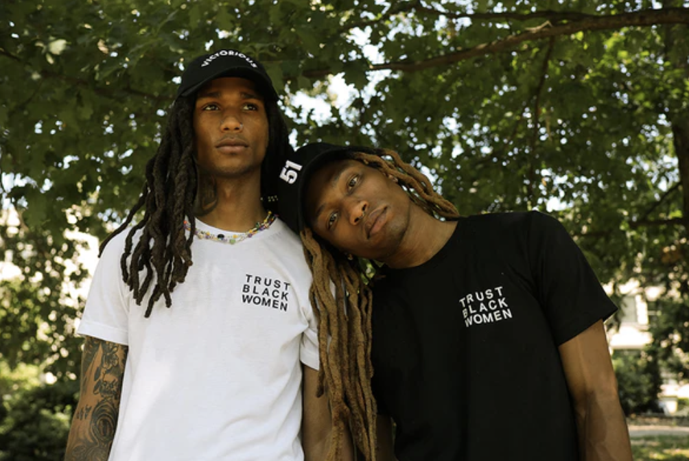 Two young Black men with dreadlocks pose outdoors under a tree, one wearing a white T-shirt and the other wearing a black T-shirt, both with 'Trust Black Women' written on them, leaning on each other.