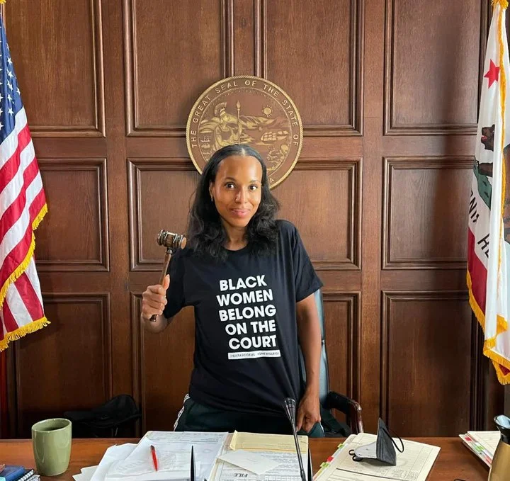 Woman holding a gavel in a courtroom, standing behind a desk with papers, wearing a black T-shirt that says 'Black Women Belong on the Court', with American and California flags in the background.