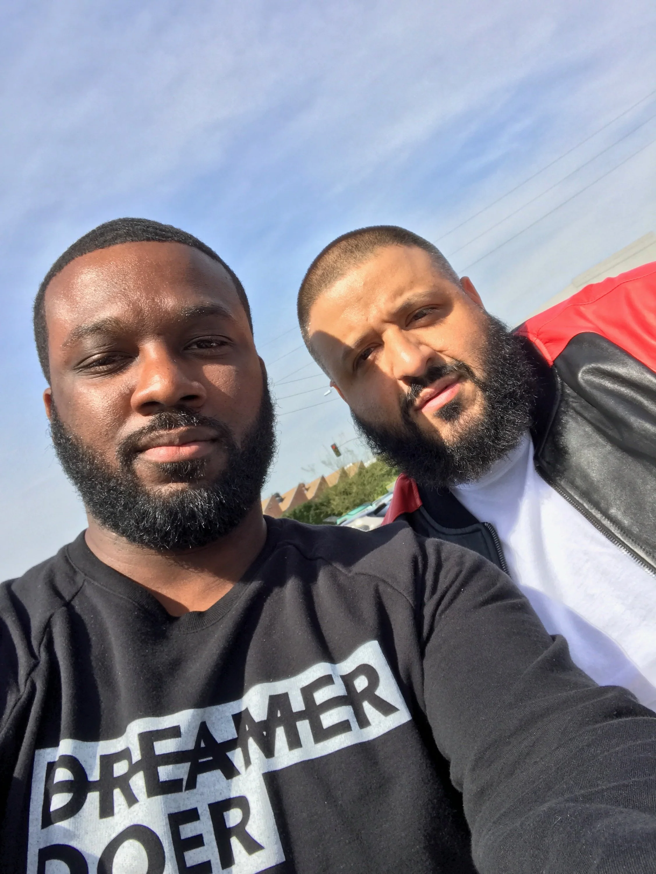 Close-up photo of two men with beards, one with a black shirt and the other with a red and black jacket, taking a selfie outdoors under a blue sky.