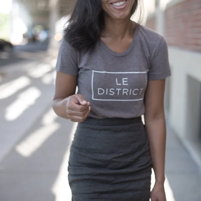 A woman walking outdoors, smiling, wearing a gray T-shirt with 'LE DISTRICT' printed on it and a dark skirt.