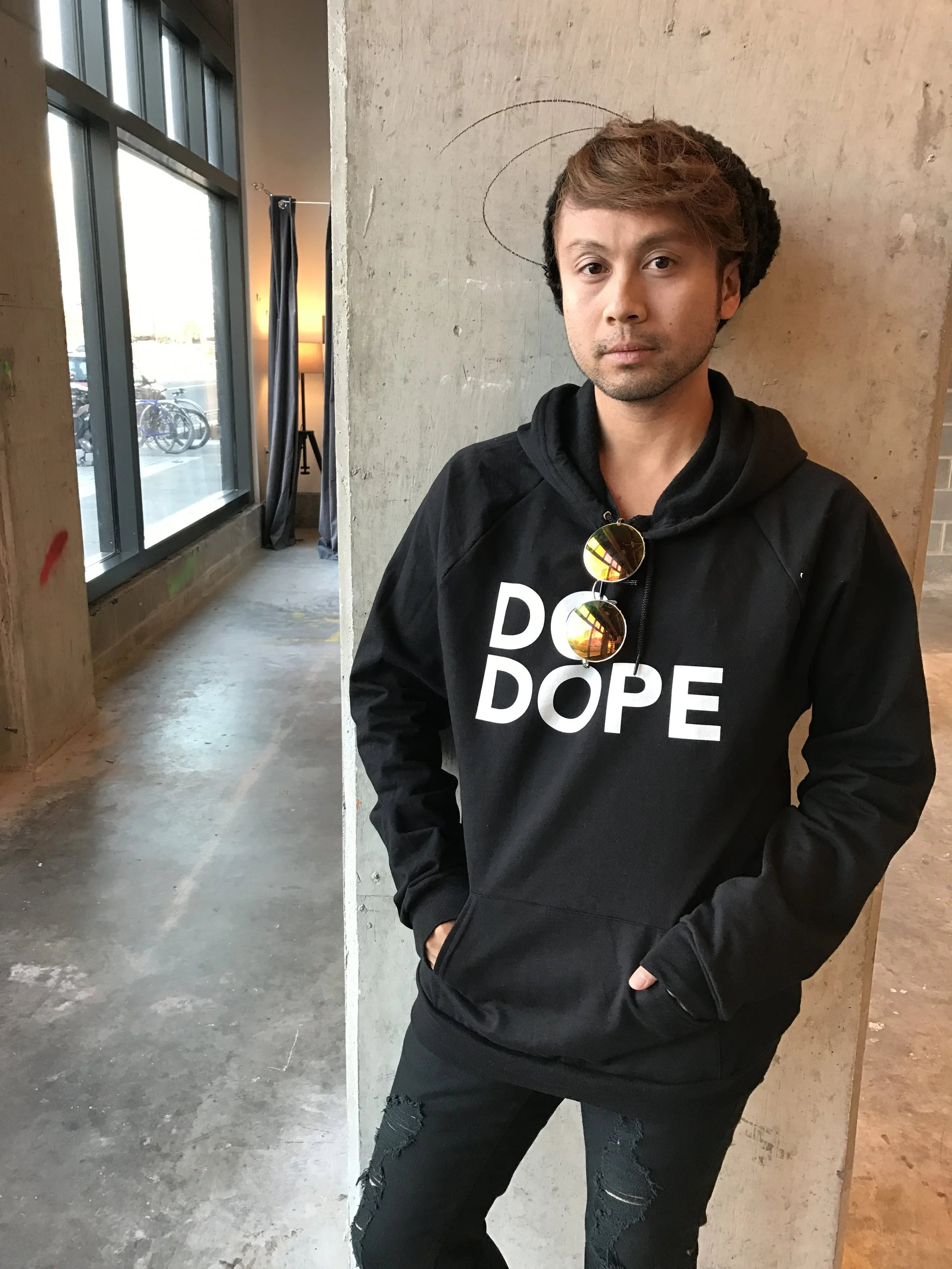 Young man with short brown hair, wearing a black hoodie with white text and ripped black jeans, leaning against a concrete wall inside a modern building with large window, curtain, and bicycle outside.