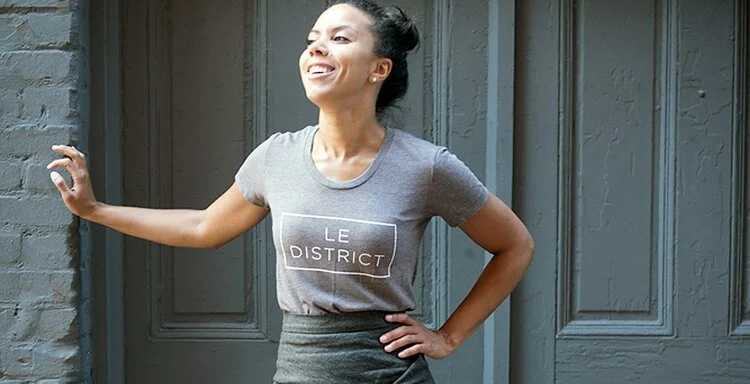 A young woman smiling and leaning against a wall outside, wearing a gray t-shirt that says "LE DISTRICT" in white letters.