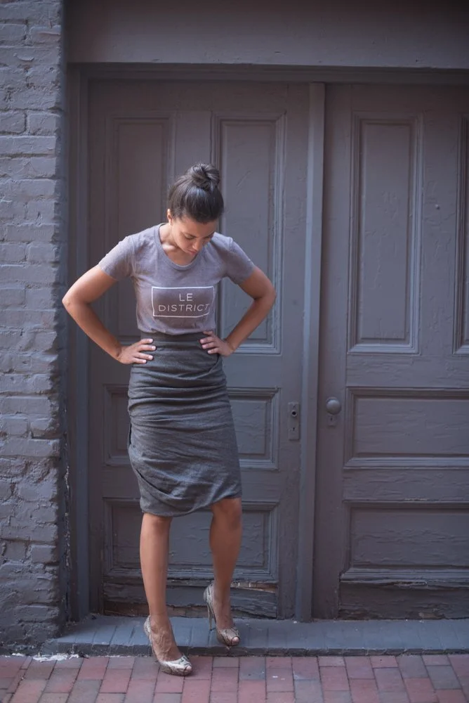 A woman standing on brick sidewalk in front of gray wooden door and brick wall, wearing a gray t-shirt with the text 'LE DISTRICT' and a gray pencil skirt, looking downward, hands on hips, wearing high heels.