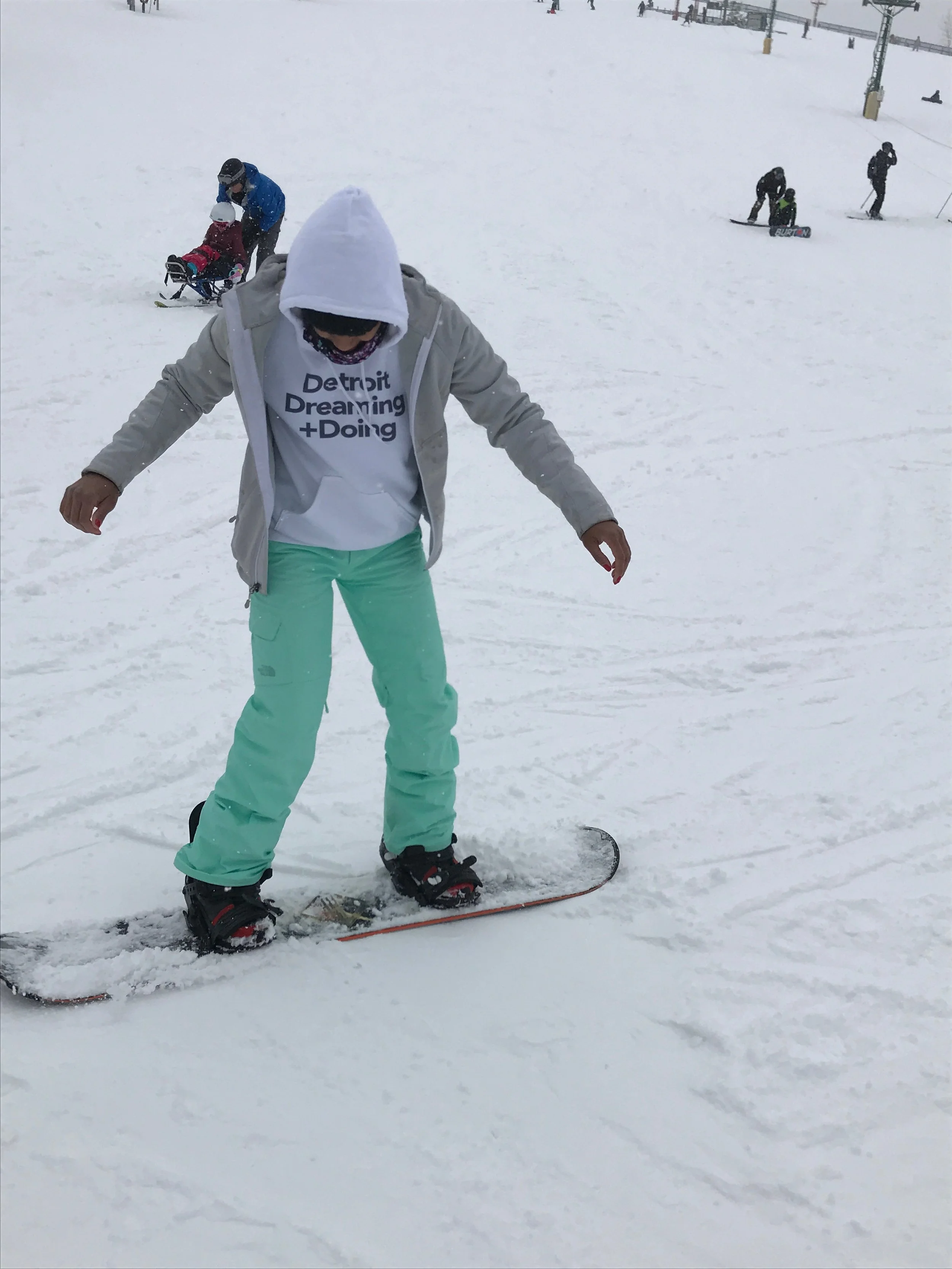 Person in a white hoodie and mint green snow pants learning to snowboard on a snowy slope, with others skiing and snowboarding in the background.