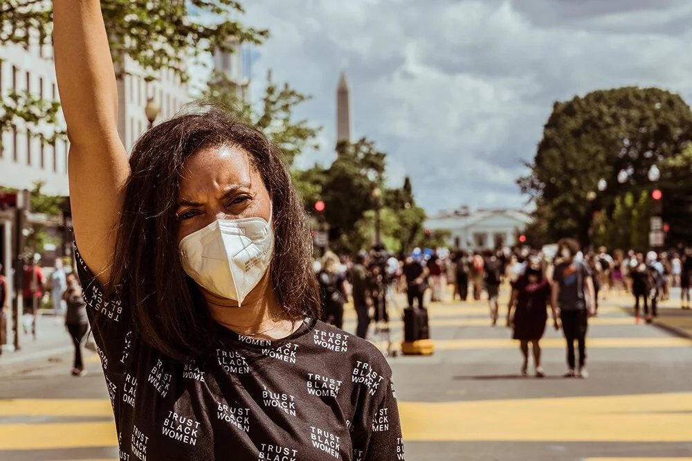 A woman wearing a face mask and a black shirt with white text, standing with her arm raised on a crosswalk in an urban area, with a crowd of people and the Washington Monument in the background.