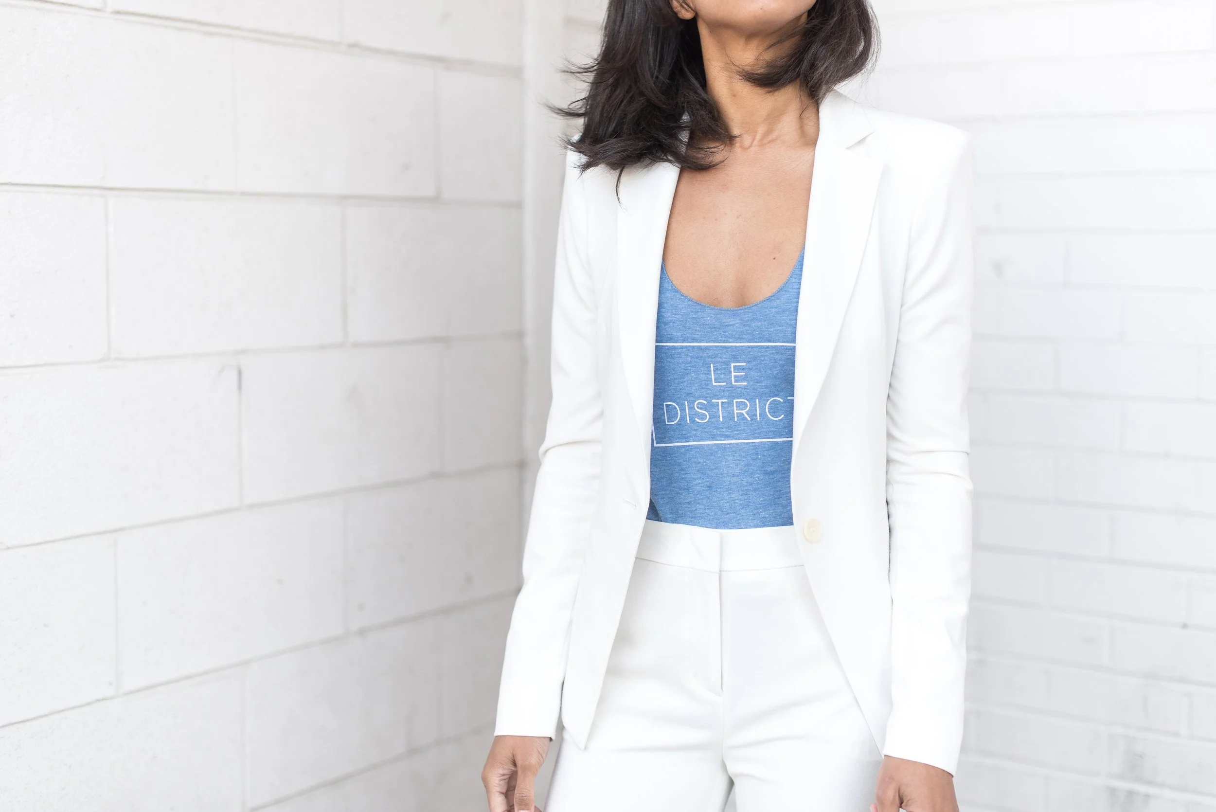 A woman wearing a white blazer and white pants, with a blue T-shirt underneath, standing against a white brick wall.