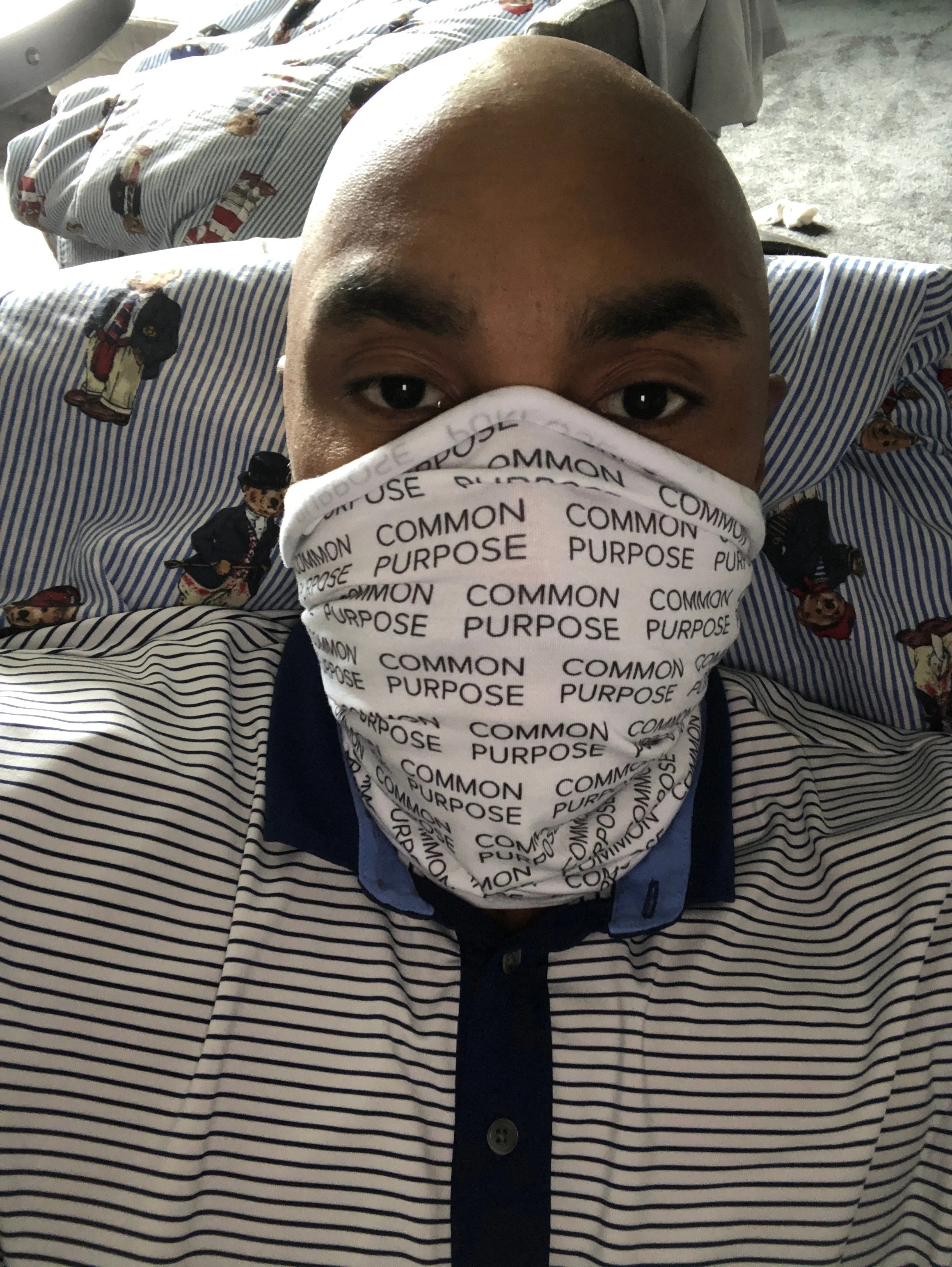 A man with a shaved head lying in bed with striped sheets featuring cartoon characters, wearing a white face mask with the words 'COMMON PURPOSE' printed repeatedly.