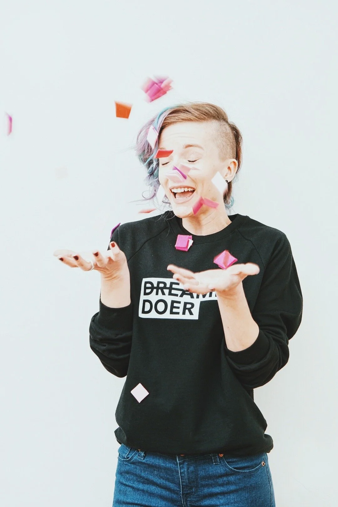 A woman with multicolored hair and a black sweatshirt with the words 'DREAM DOER' celebrates with falling confetti.
