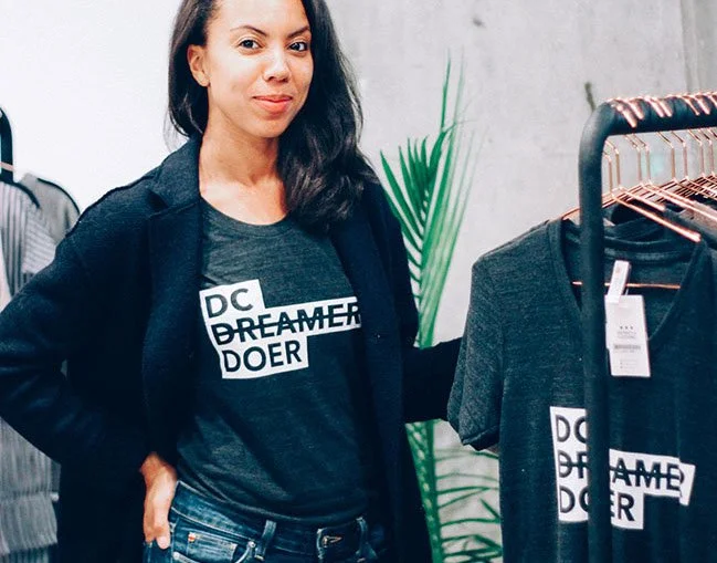 A woman shopping for clothes, standing next to a rack of dark-colored t-shirts with the words 'DC DREAMER DOER' printed on them, in a retail store with a gray wall and a potted plant in the background.
