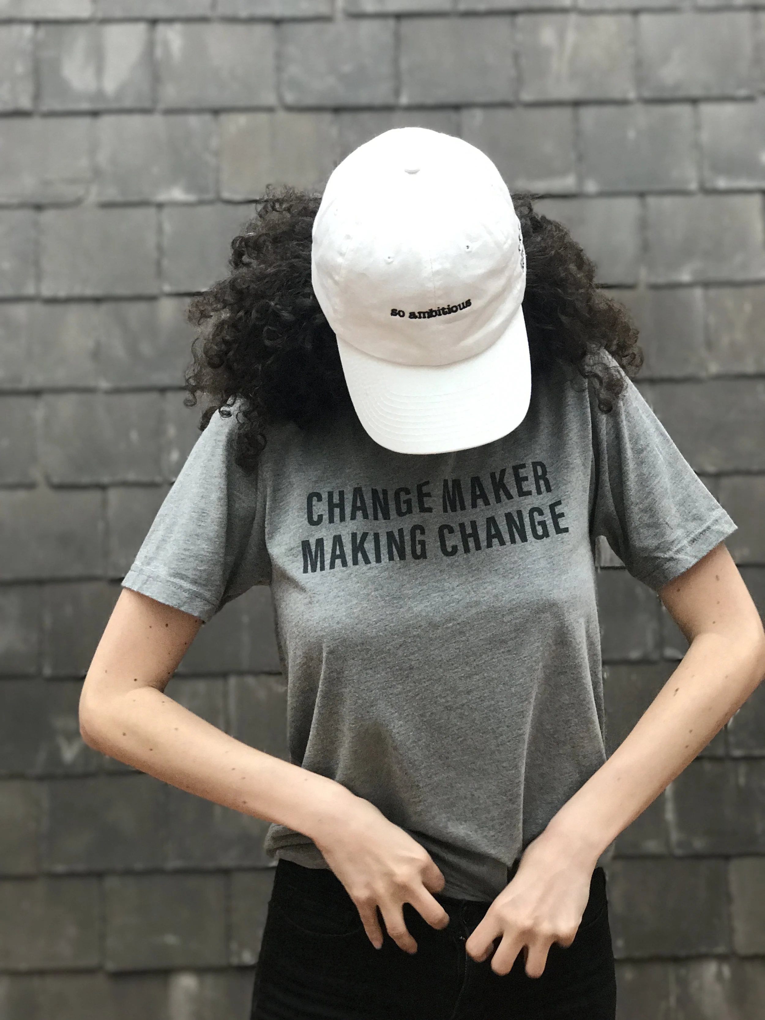 Person wearing a white cap with the words 'so ambitious' embroidered on it, standing in front of a dark gray brick wall, wearing a gray T-shirt with words 'CHANGE MAKER' printed on it, with one hand near their waist and the other arm crossed.