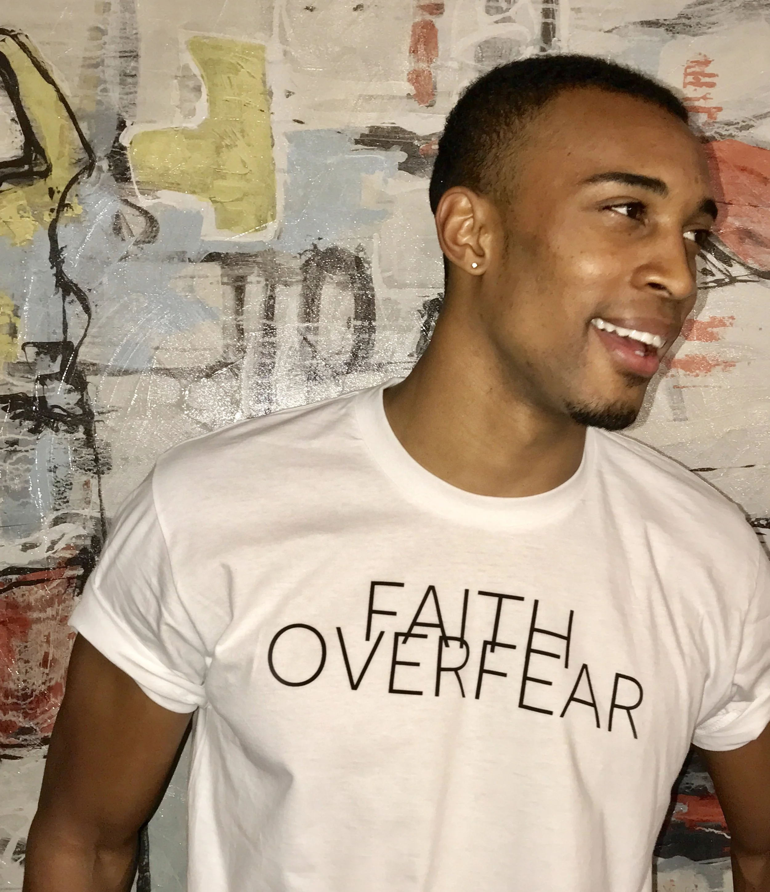 Close-up of a smiling young man with short hair and a small earring, wearing a white t-shirt with black text, standing in front of an abstract painting with various colors and textures.