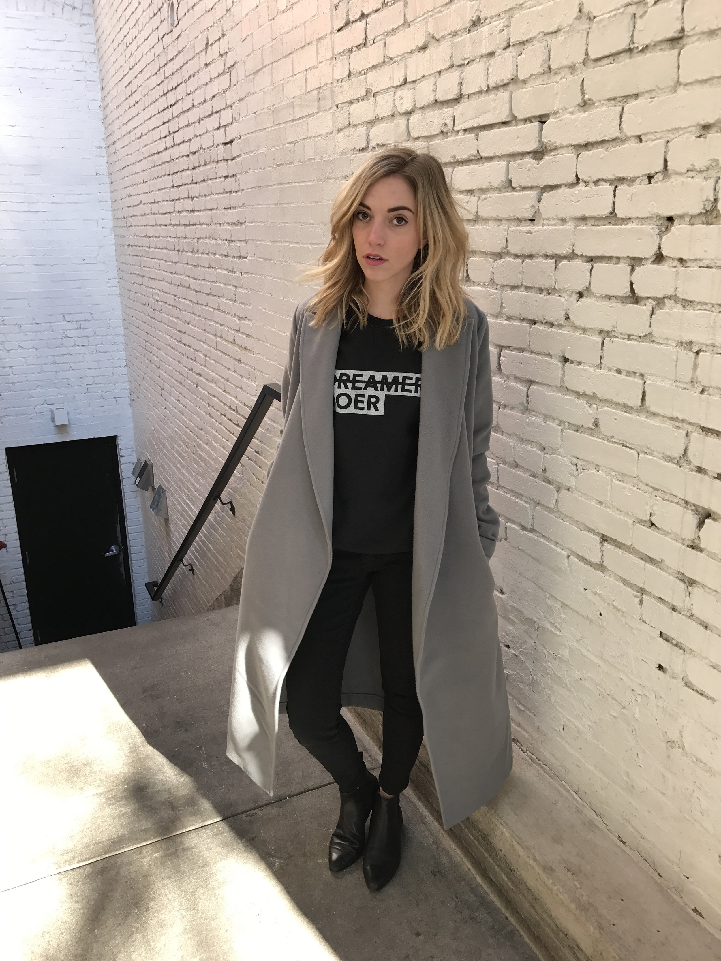 A woman with blonde wavy hair standing against a white brick wall. She is wearing a black T-shirt with the words 'DREAMER DOER' in white text, a long gray coat, black pants, and black boots. She is looking at the camera with a neutral expression.