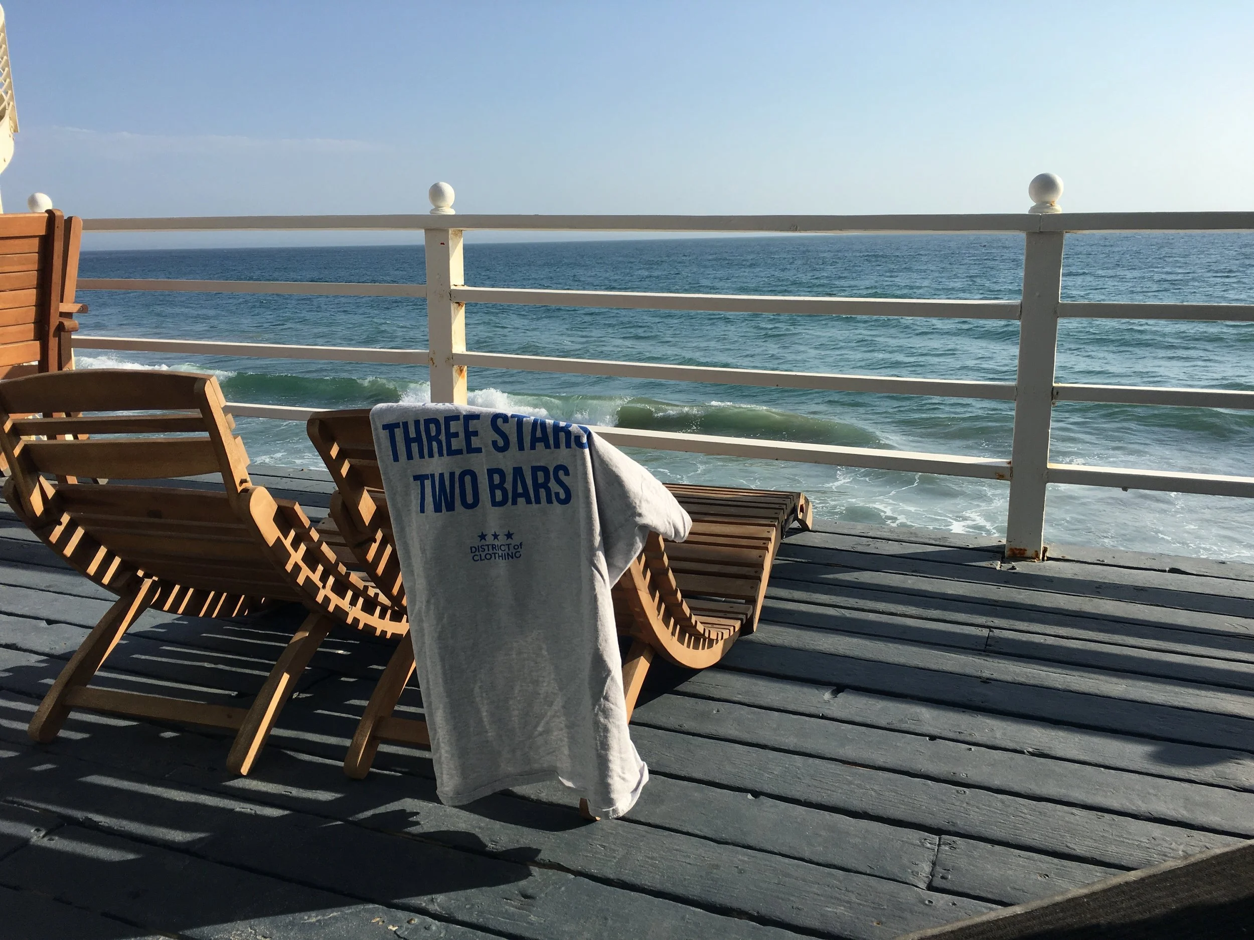 View of the ocean from a wooden deck with wooden chairs, a white railing, and a towel with blue text hanging over a chair, under a clear sky.