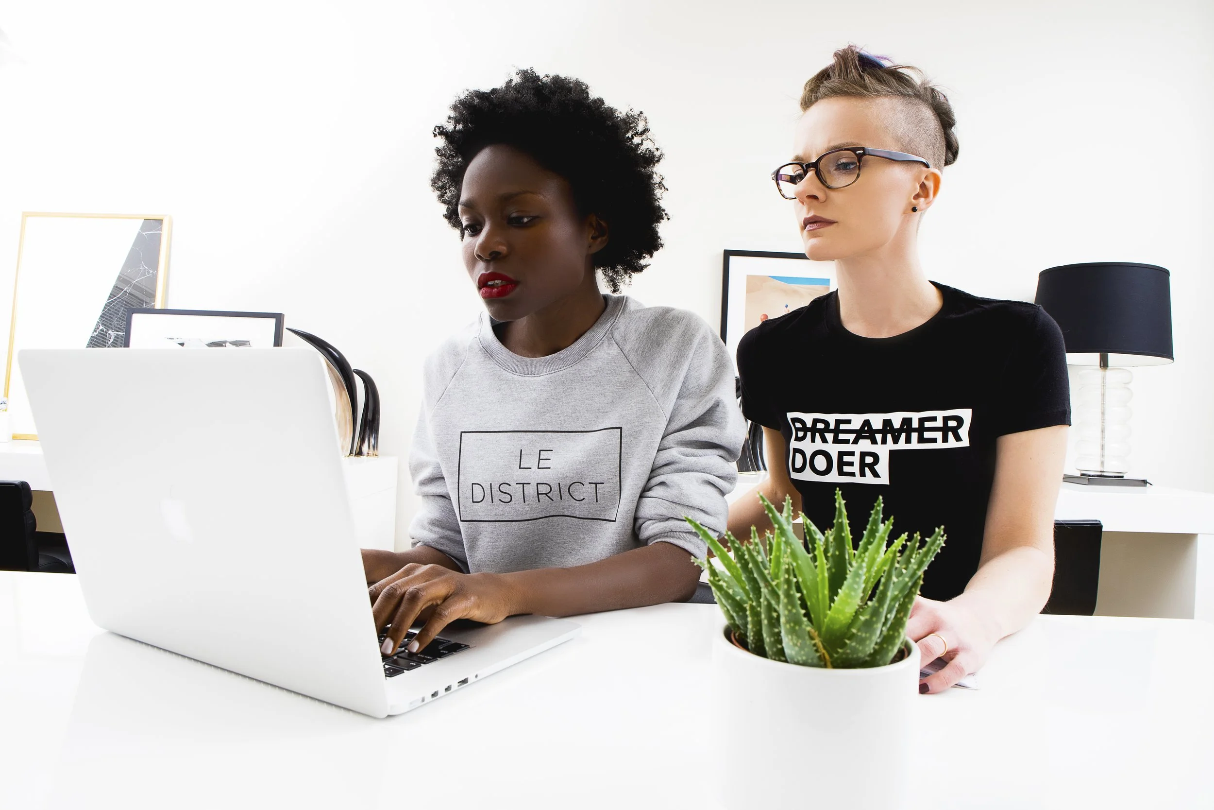 Two women working together at a white desk with a silver laptop and potted cactus plant, in a bright room with minimal decor.