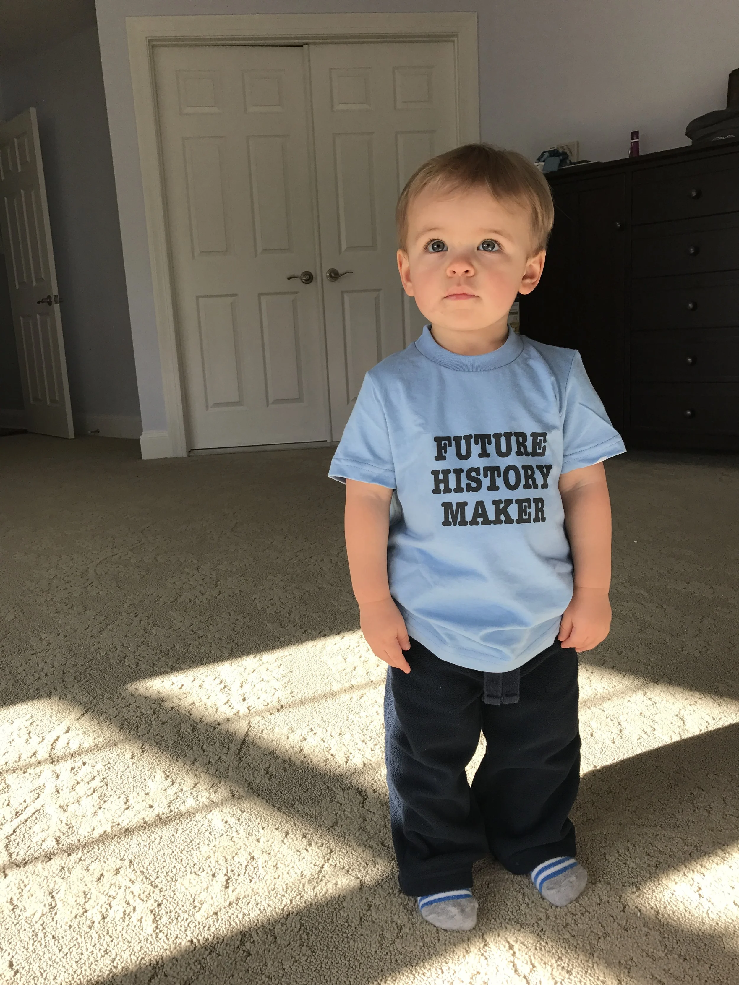 A young boy standing indoors on a carpeted floor, wearing a light blue shirt with bold black text that says 'FUTURE HISTORY MAKER,' black pants, and striped socks. The background features closed white double doors and a dark dresser.