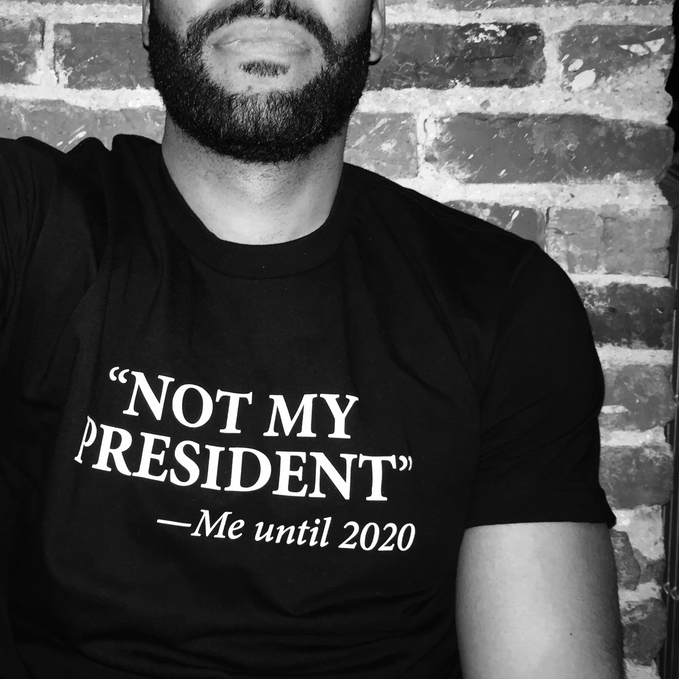 Close-up black and white photo of a person's face, focusing on the beard and mouth, wearing a black T-shirt with white text, against a brick wall background.