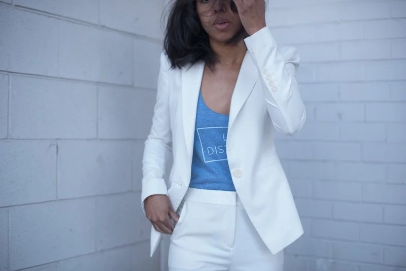 A woman with dark hair wearing a white suit over a blue graphic T-shirt stands against a light gray brick wall, partially covering her face with her hand.