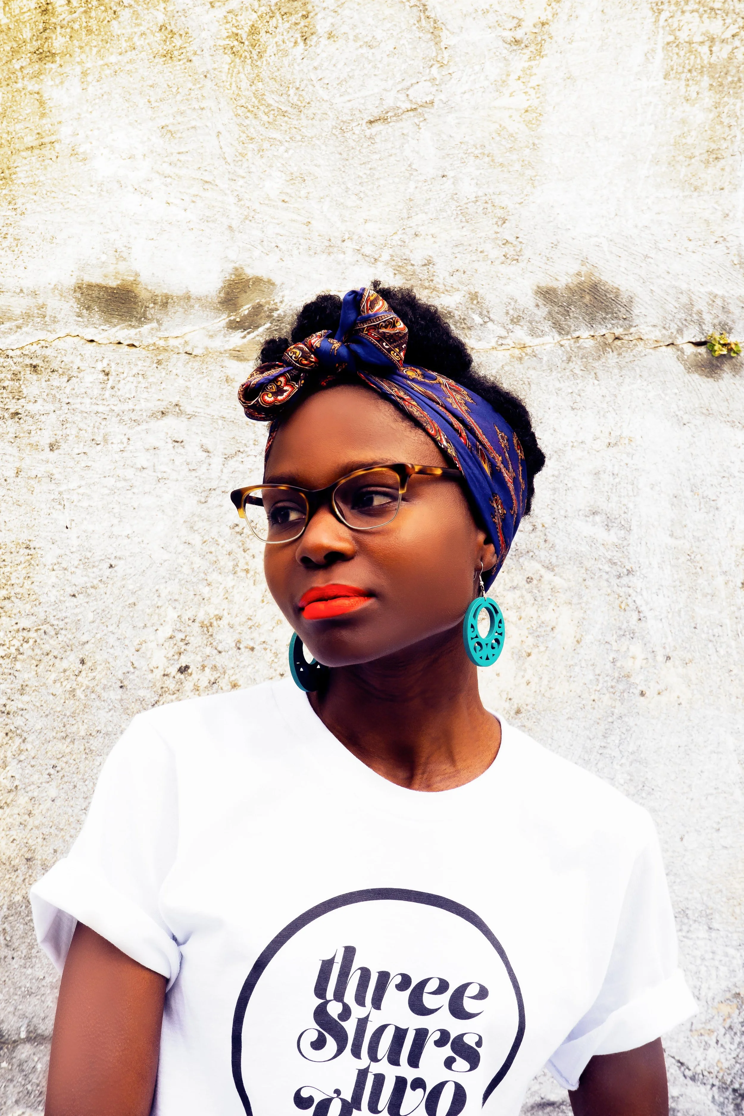 A woman with dark curly hair and glasses, wearing a blue headscarf with colorful patterns, large turquoise earrings, and a white T-shirt with the words 'three stars' on it, standing in front of a textured, off-white wall.
