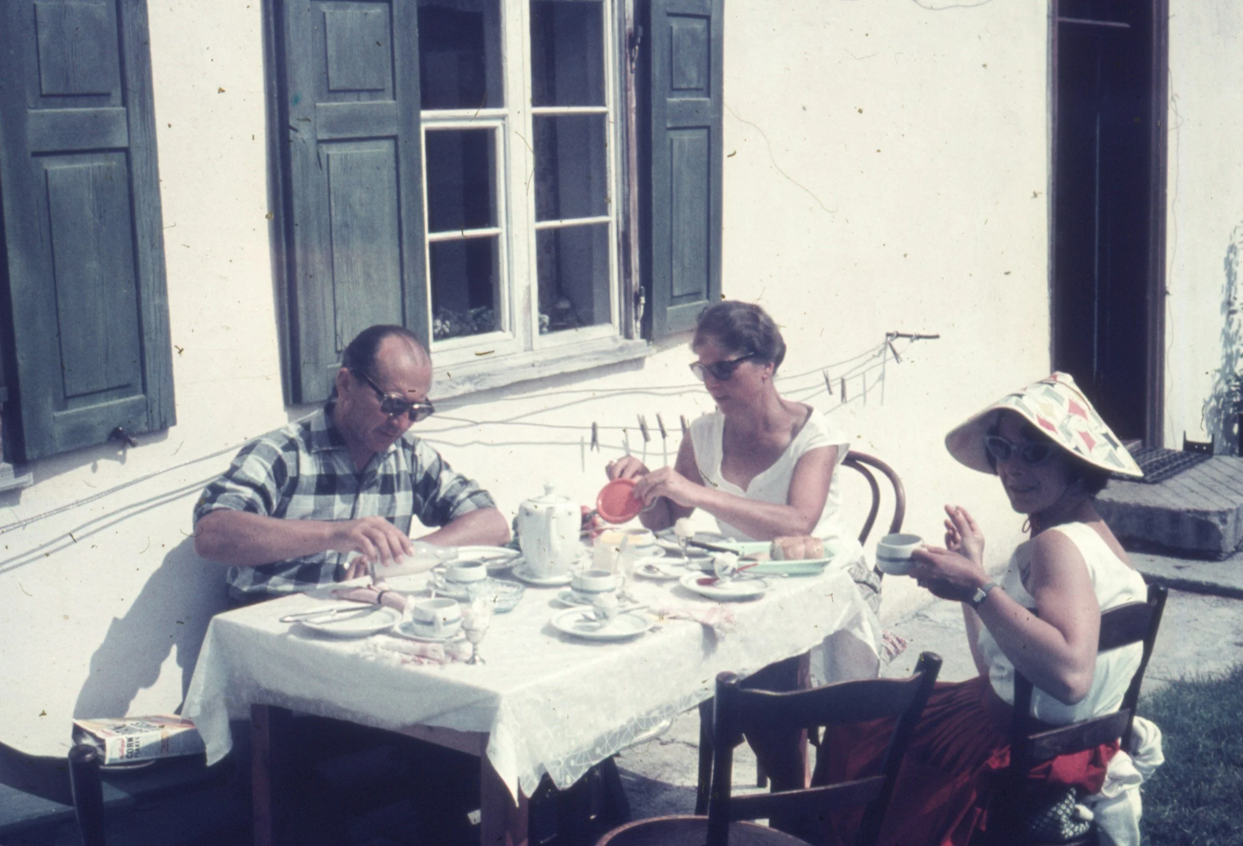Three people sitting at a table outdoors, enjoying a meal and drinking tea, with a window and shutters in the background.