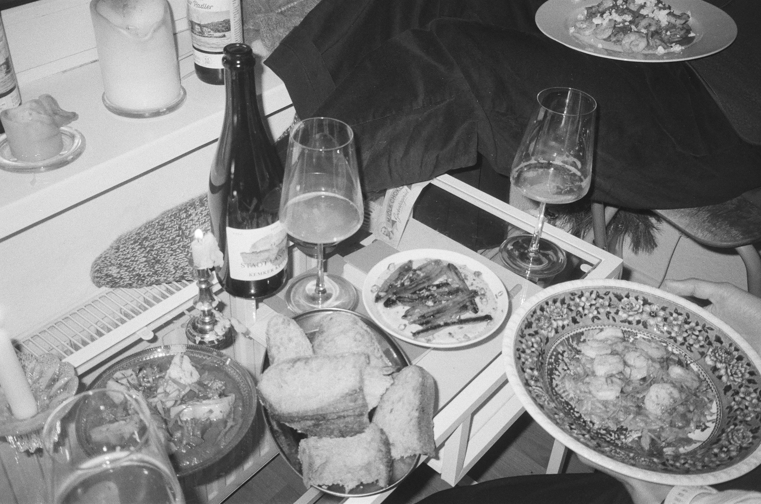 A table filled with various dishes of food, glasses of wine, and bottles. The table has a striped tablecloth and there are multiple plates of food, including a pizza, a seafood dish, and salads. There are also candles and a person wearing dark clothing sitting at the table.