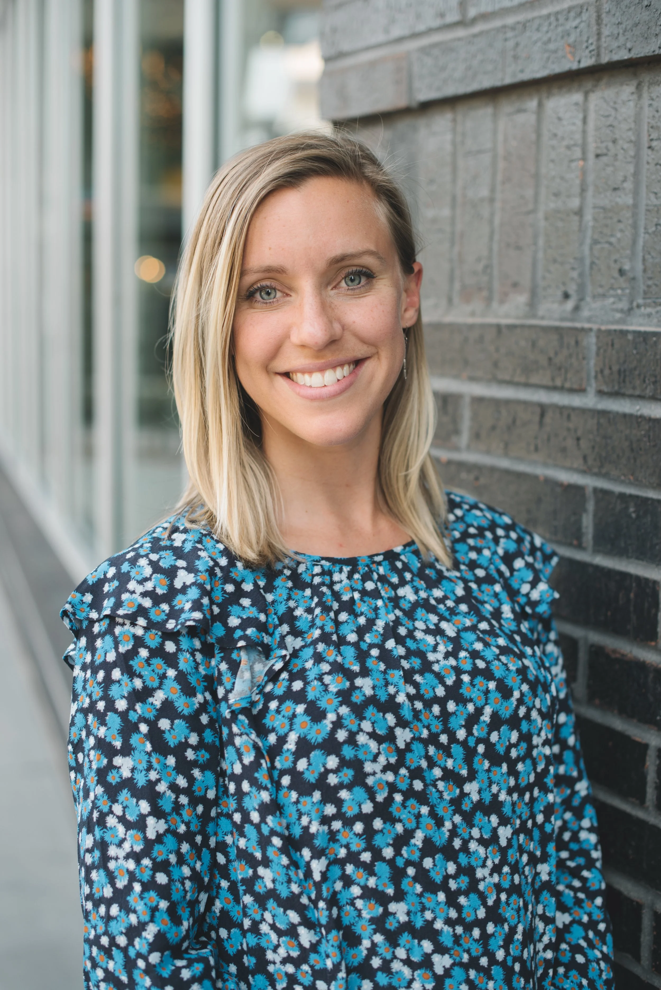 A young woman with blonde hair and blue eyes smiling at the camera, standing outdoors against a brick wall, wearing a blue floral dress.