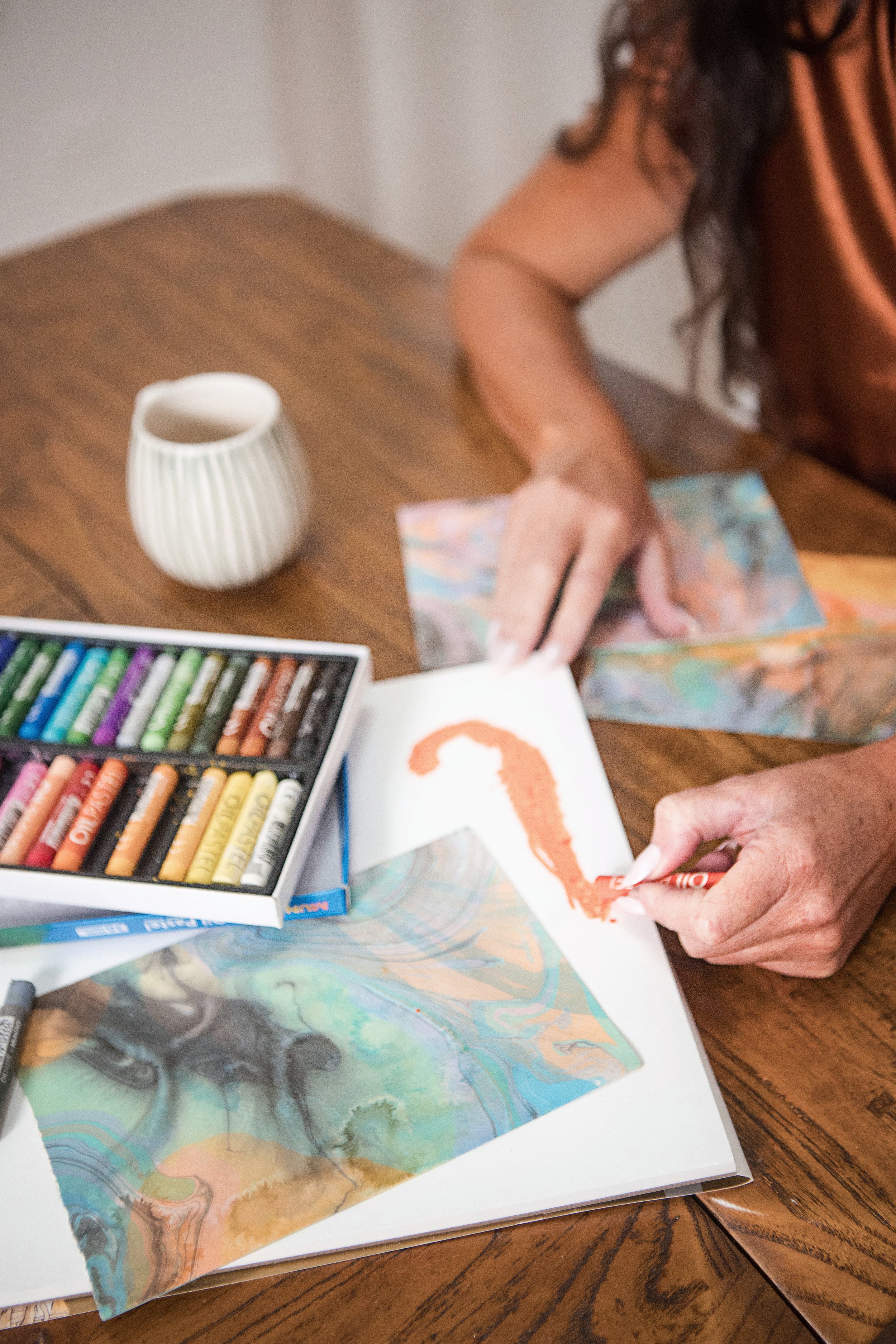 Close up of oil pastels, layered art work and Kylie's hands using an orange pastel during a creative process.