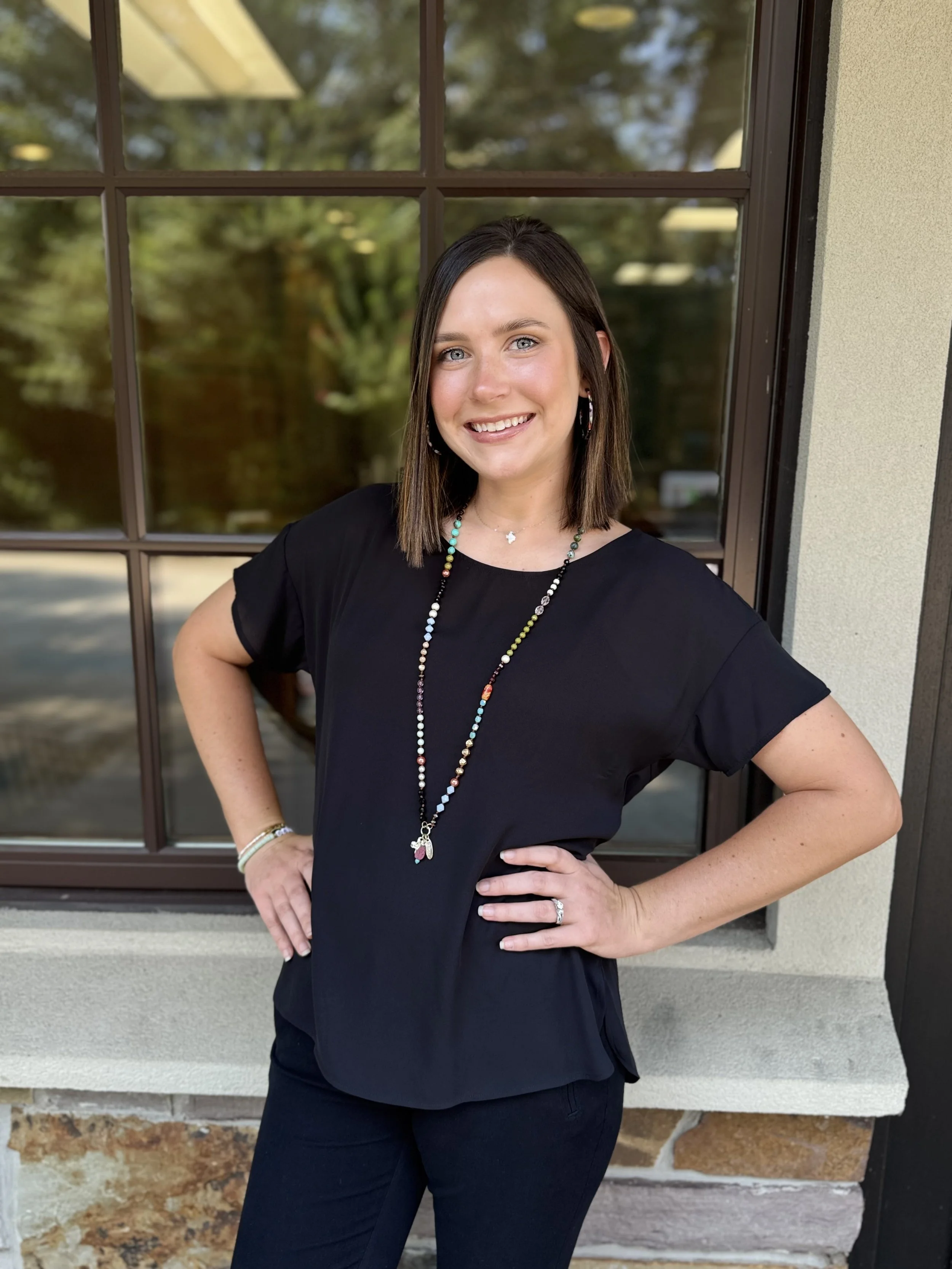 A woman with shoulder-length dark hair, wearing a black short-sleeve top and black pants, standing outside near a window with a glass pane and beige wall, smiling with her hands on her hips, wearing earrings, a necklace, a ring, and a bracelet.