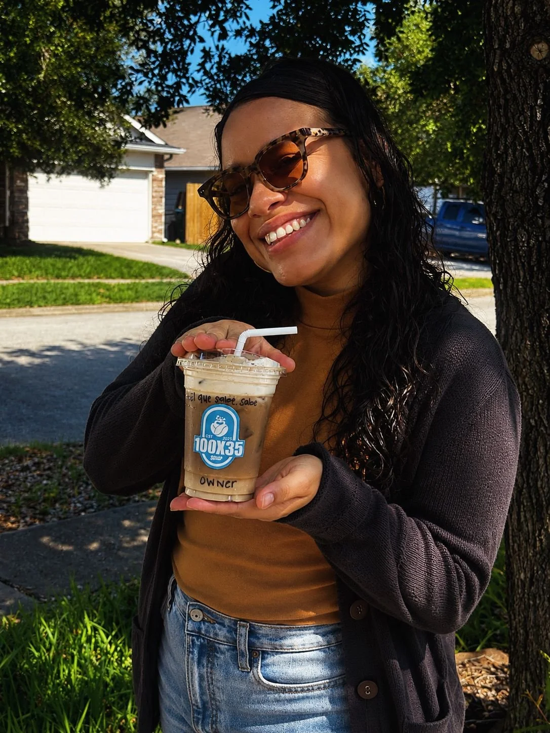 A woman with curly hair wearing sunglasses and a black cardigan smiling outdoors while holding a plastic cup of iced coffee with a straw.