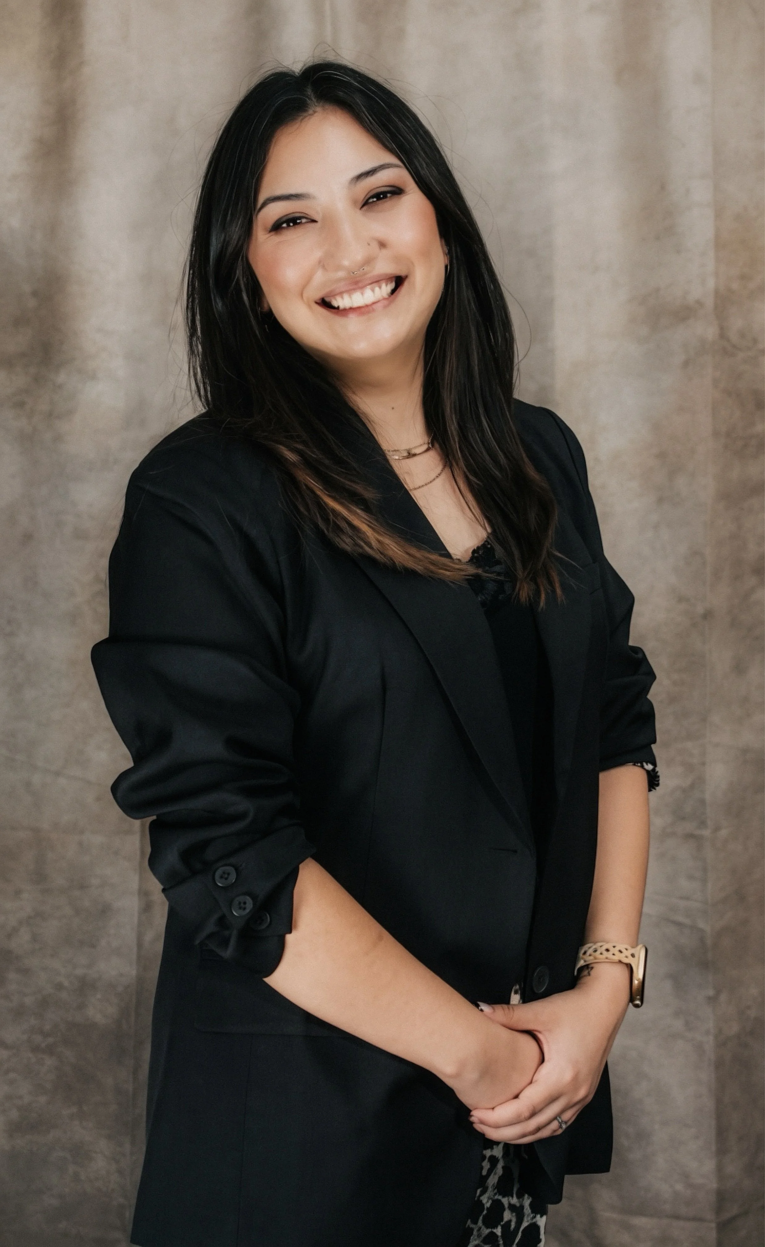 A young woman with long black hair, smiling and wearing a black blazer, a watch, and a patterned dress, standing against a plain background.