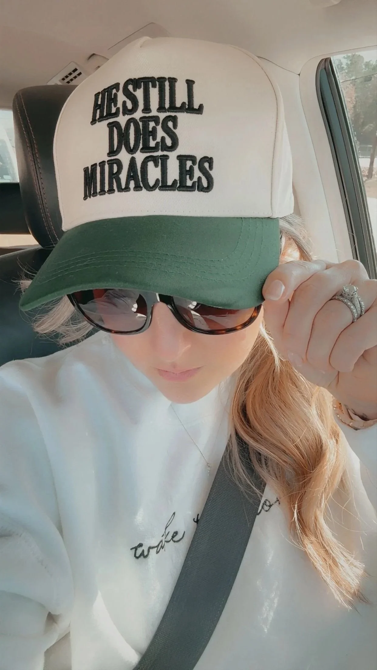A woman wearing sunglasses and a baseball cap with the quote 'Hustle Does Miracles', sitting in a car, adjusting her cap with her hand, wearing a white sweatshirt.