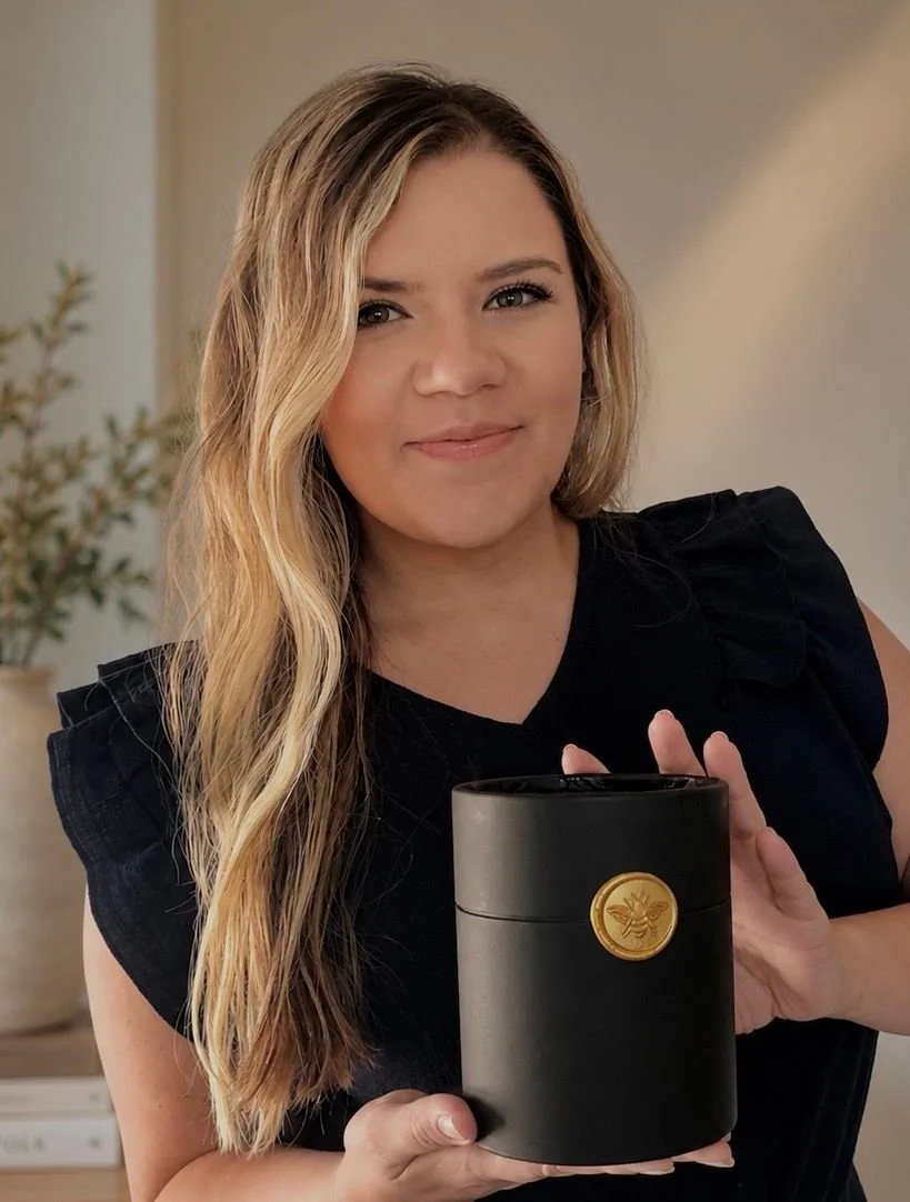 A woman with long blonde wavy hair smiling and holding a black box with a gold emblem on it in an indoor setting.