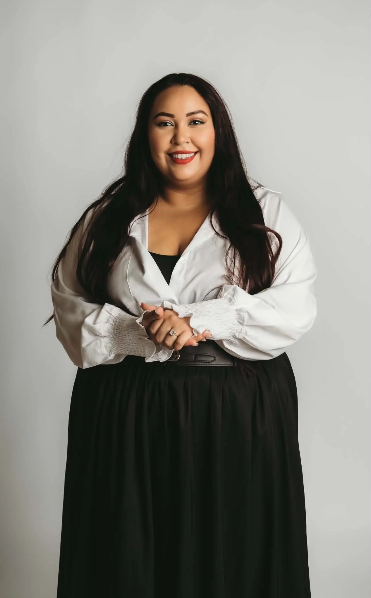 A woman with long dark hair smiling, wearing a white blouse and black high-waisted skirt, standing against a plain gray background.