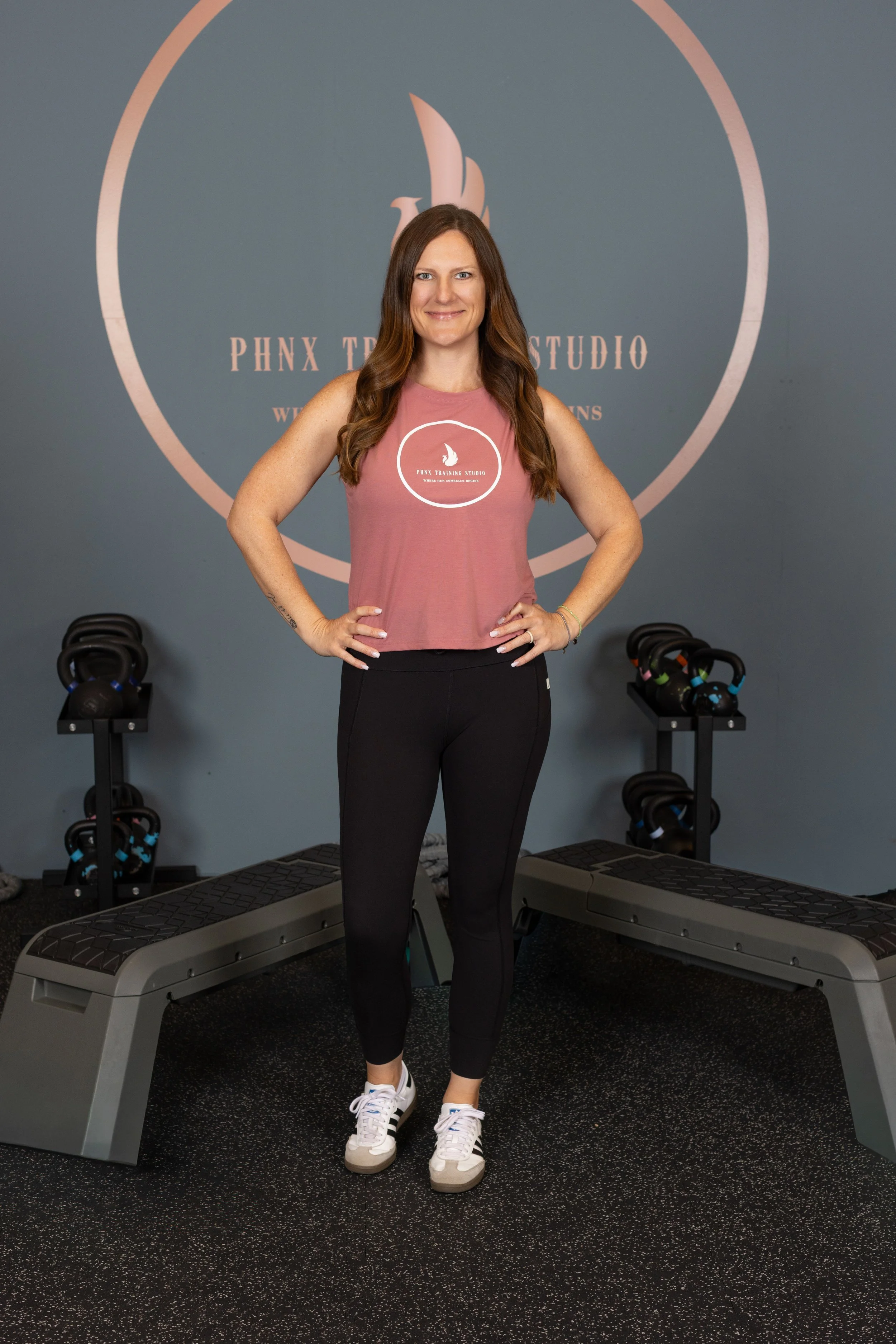 A woman standing with hands on hips in a gym fitness studio, wearing a pink sleeveless top and black leggings, behind her are kettlebells on racks and a bench, and a wall with a logo reading 'PHNX TRAINING STUDIO'.