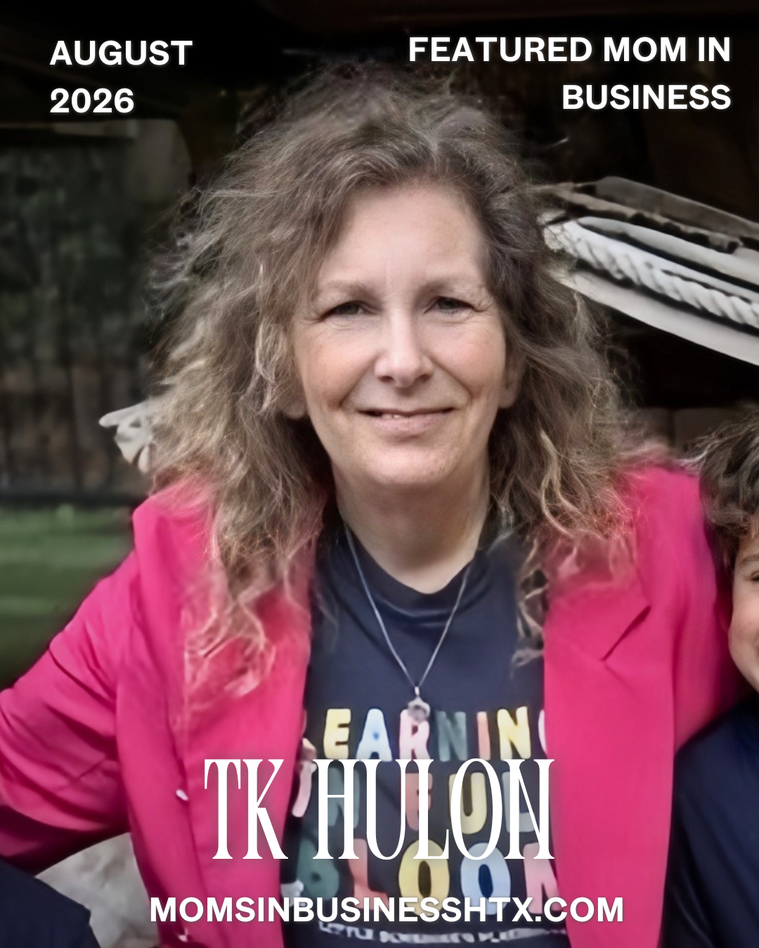 A woman with curly, shoulder-length hair smiling, wearing a pink blazer over a black shirt with colorful lettering, standing outdoors in front of a wooden structure.