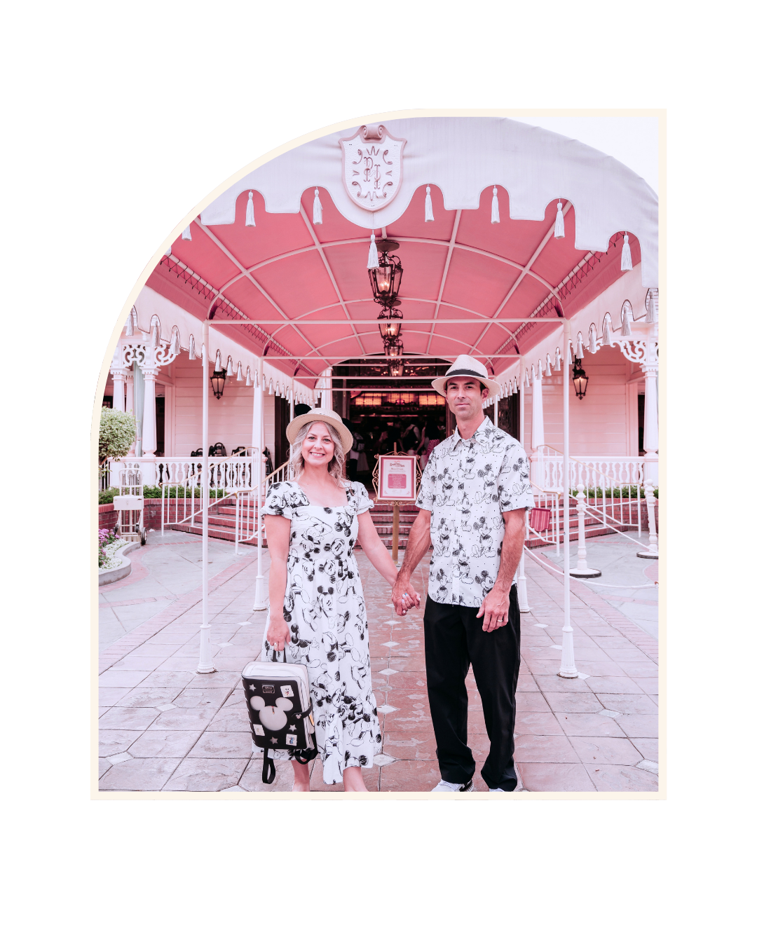 A couple standing outside a pink building, holding hands, wearing matching Mickey Mouse-themed outfits and hats.