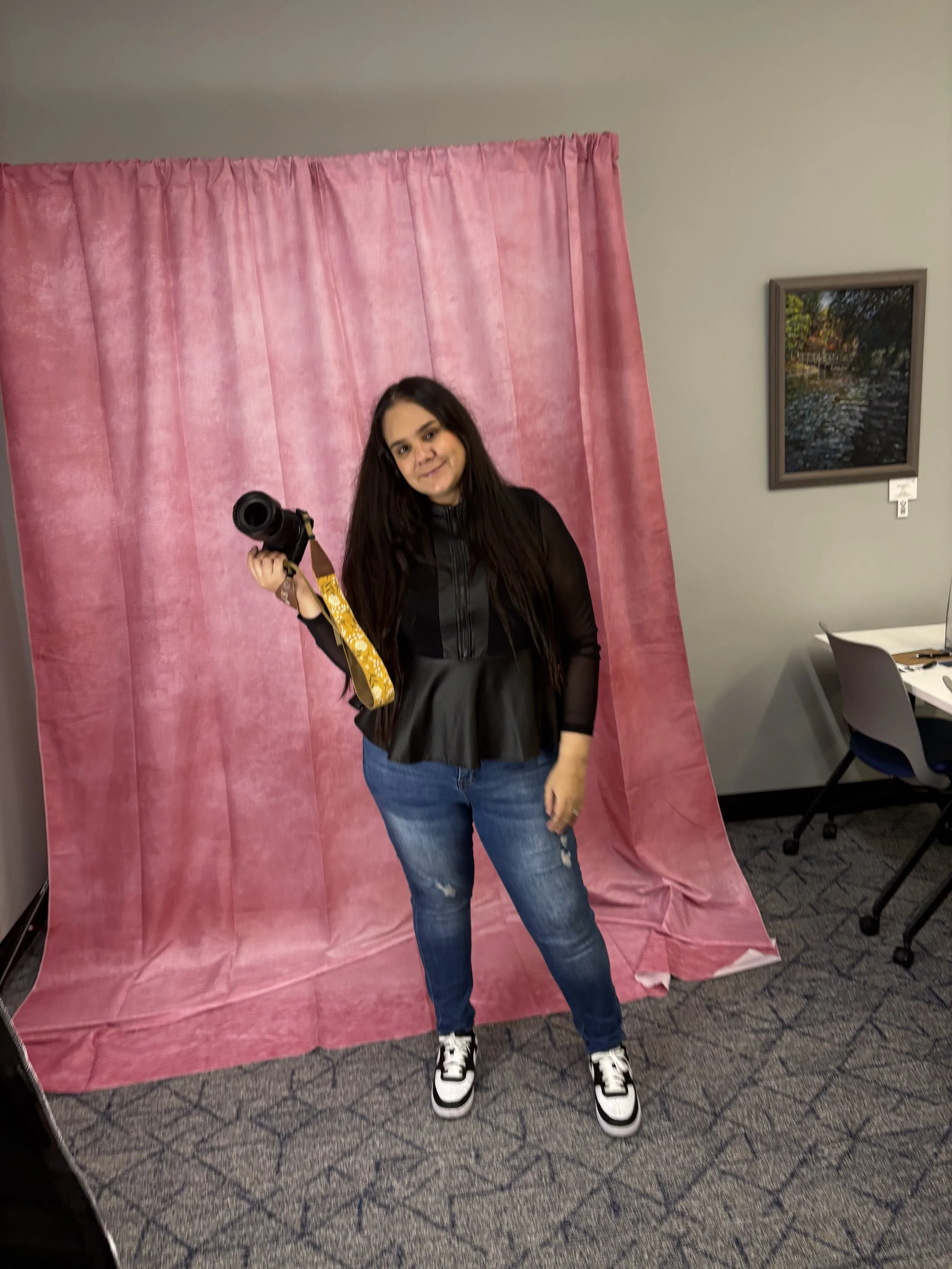 A young woman with long dark hair standing in front of a pink fabric backdrop, holding a camera with a yellow strap. She is wearing a black jacket, blue jeans, and black-and-white sneakers.
