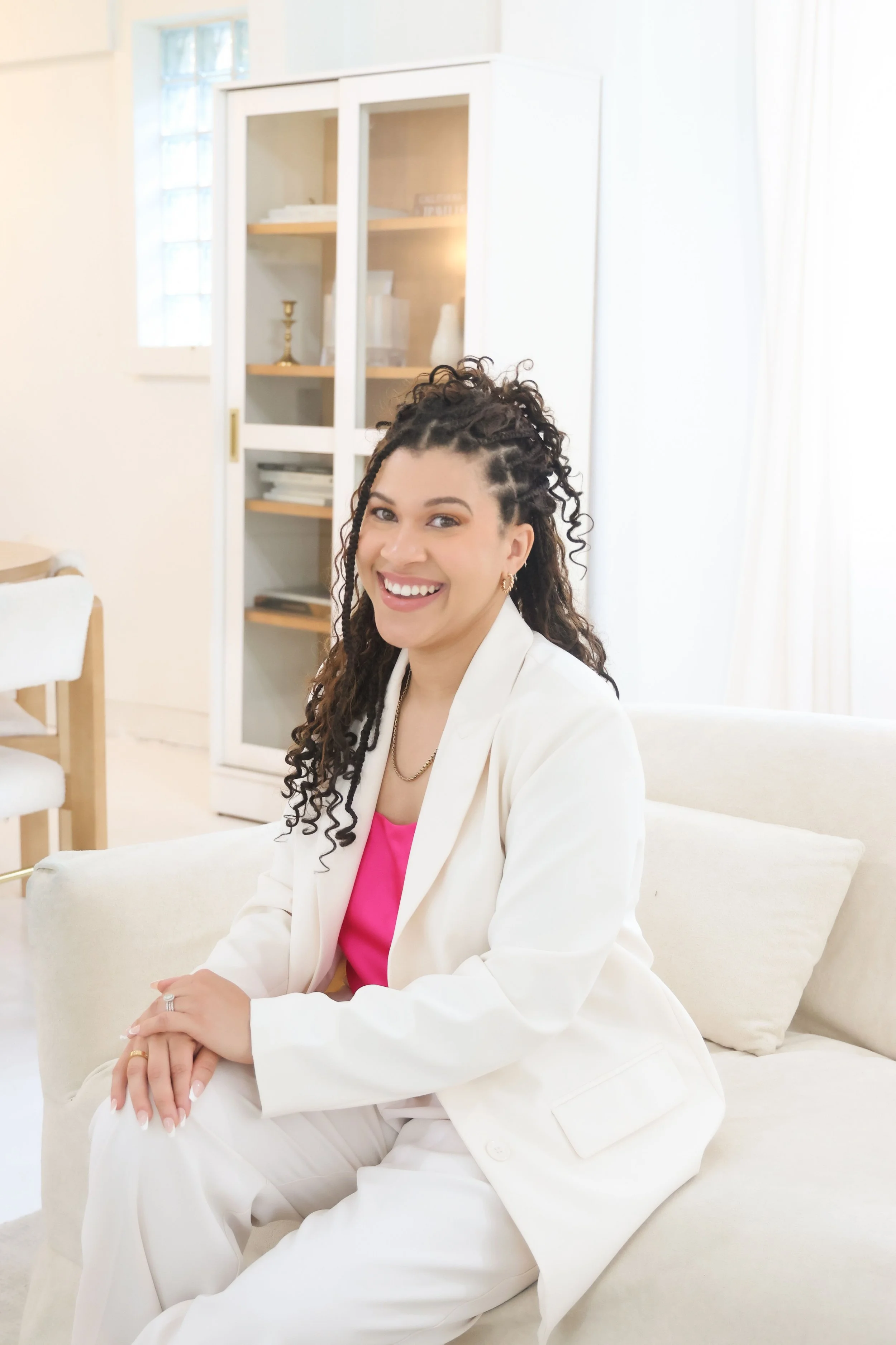 A woman with curly hair and gold jewelry sitting on a cream-colored sofa in a bright room with white walls and furniture, smiling at the camera.