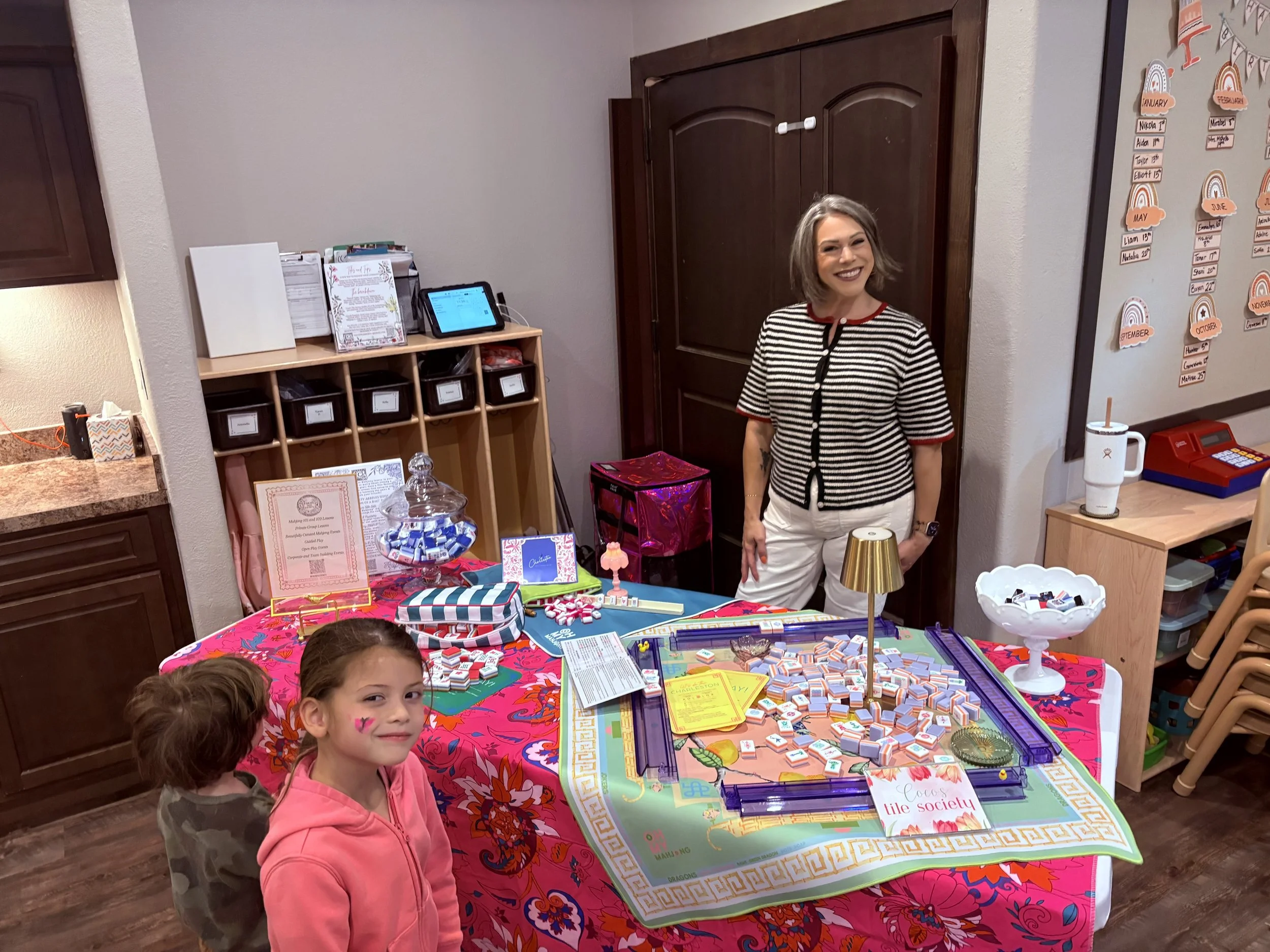 A woman smiling stands behind a table with various small objects and paper items, inside a room with a whiteboard and shelves in the background.