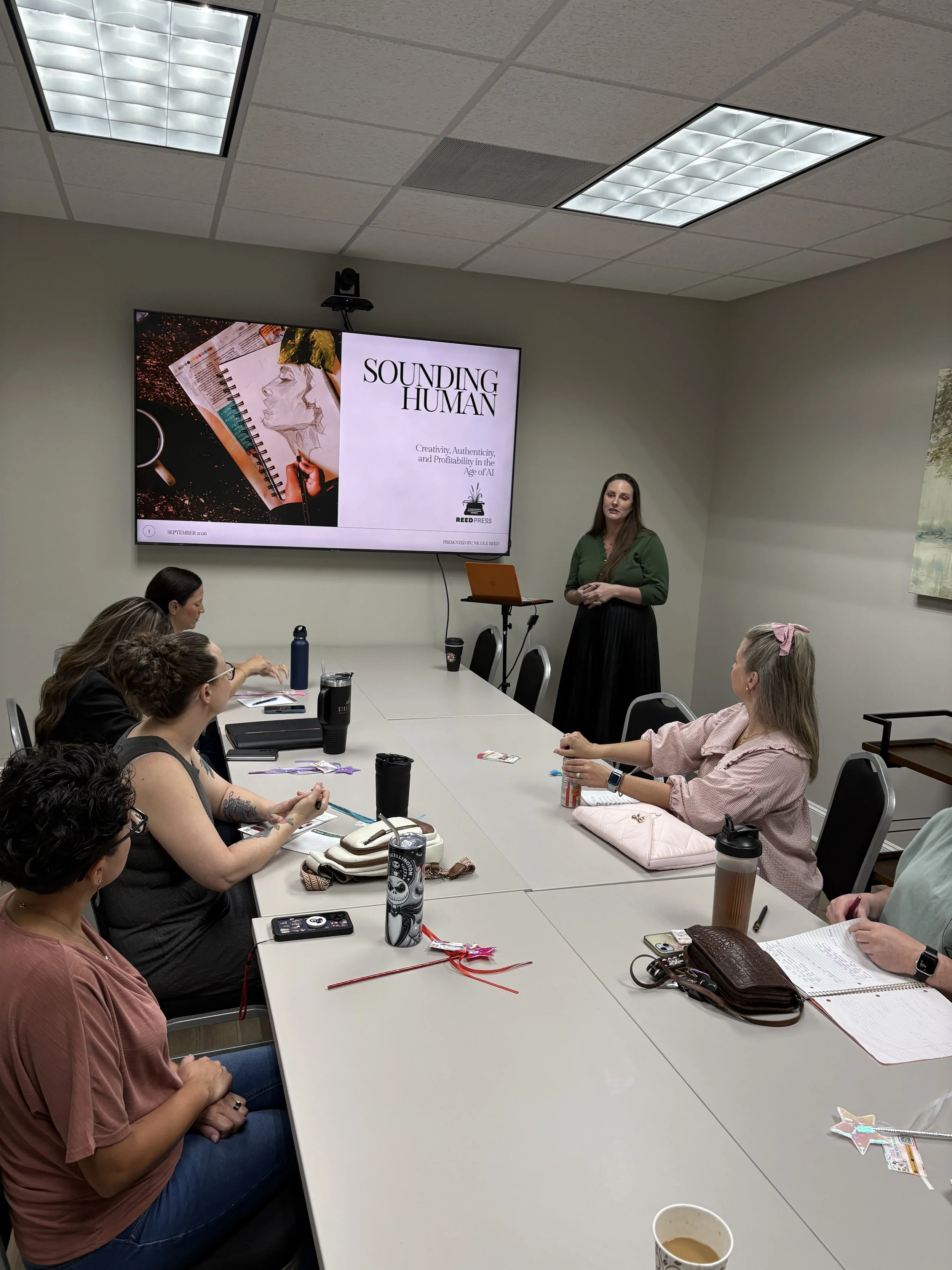 A group of women seated around a conference table in a meeting room, listening to a woman standing and speaking with a presentation slide on a large screen behind her. The slide is titled 'Sounding Human' and discusses creativity, authenticity, and p
