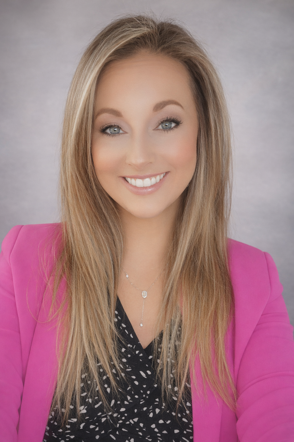 A smiling woman with long blonde hair, wearing a bright pink blazer and a black patterned blouse, with a silver necklace, against a neutral background.