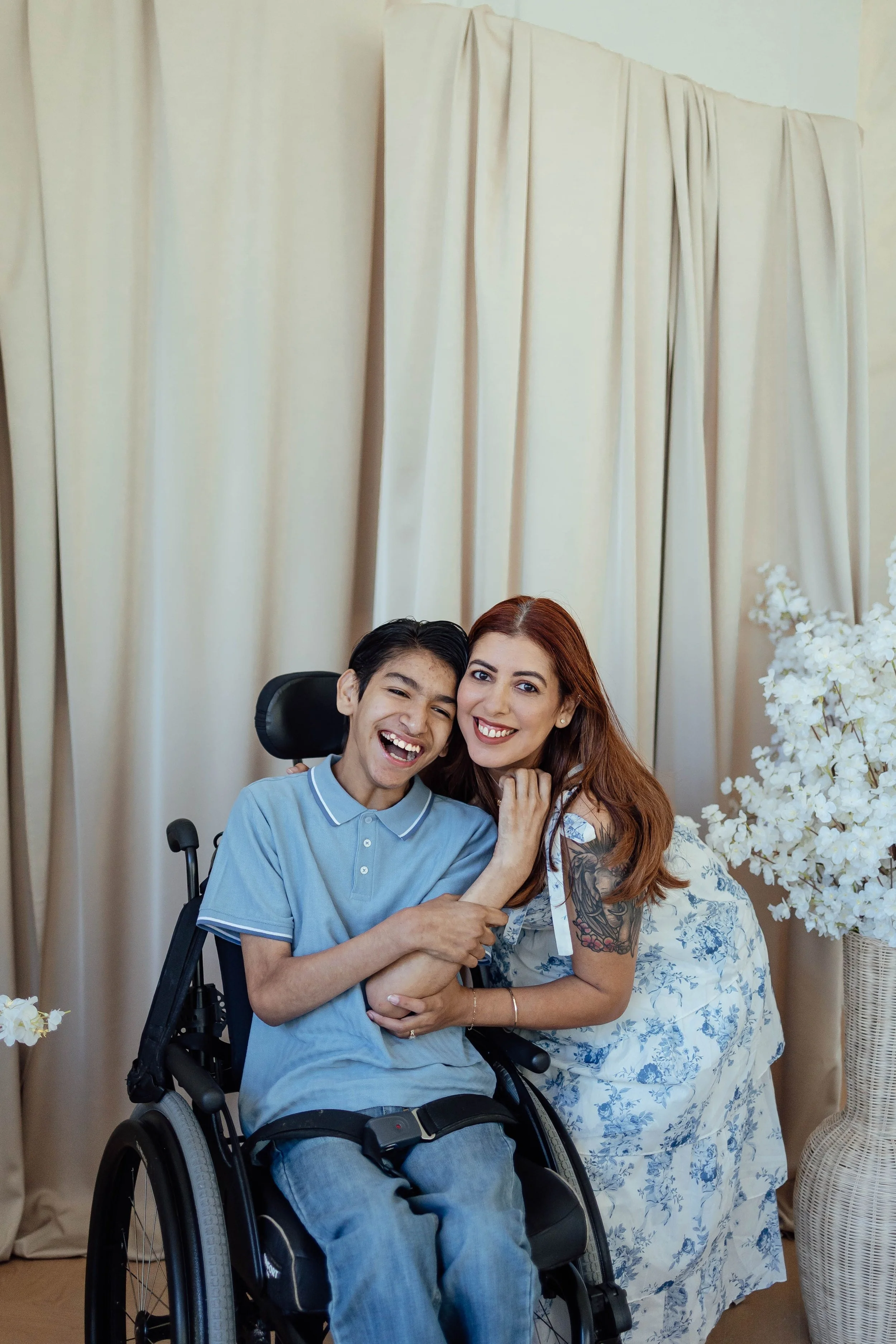 A young man with short dark hair and a woman with long red hair are smiling and hugging each other. The young man is sitting in a wheelchair. The woman has tattoos on her left arm. They are in front of beige curtains and next to a large vase with white flowers.