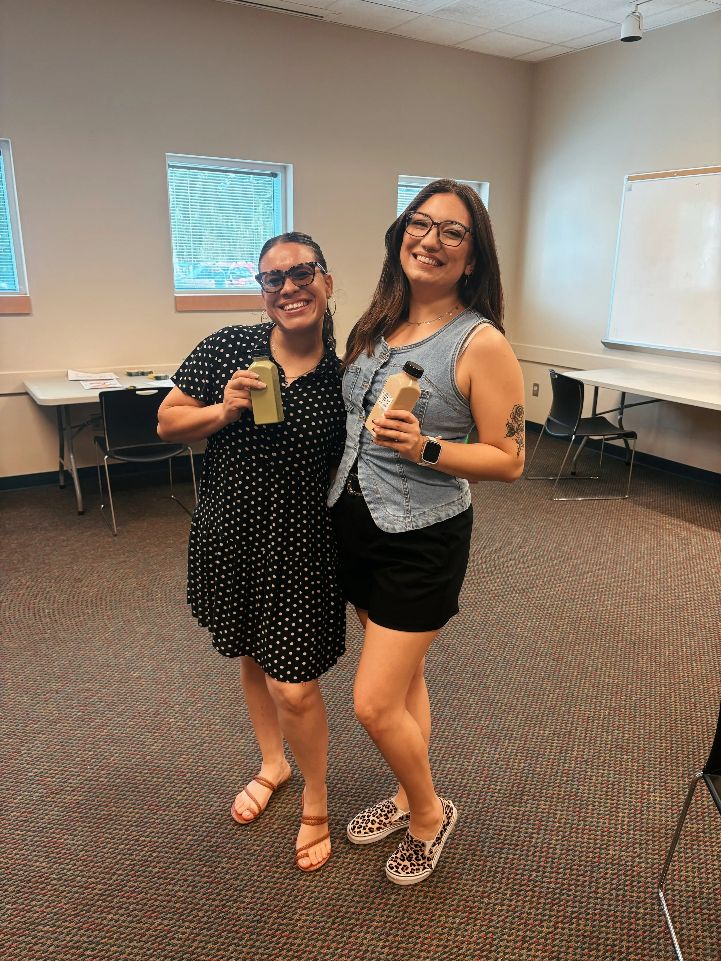 Two women standing indoors in a room with beige walls and carpet, holding drinks and smiling at the camera. One woman is wearing a black polka-dot dress, sandals, and glasses. The other woman is wearing a sleeveless denim vest, black shorts, leopard 