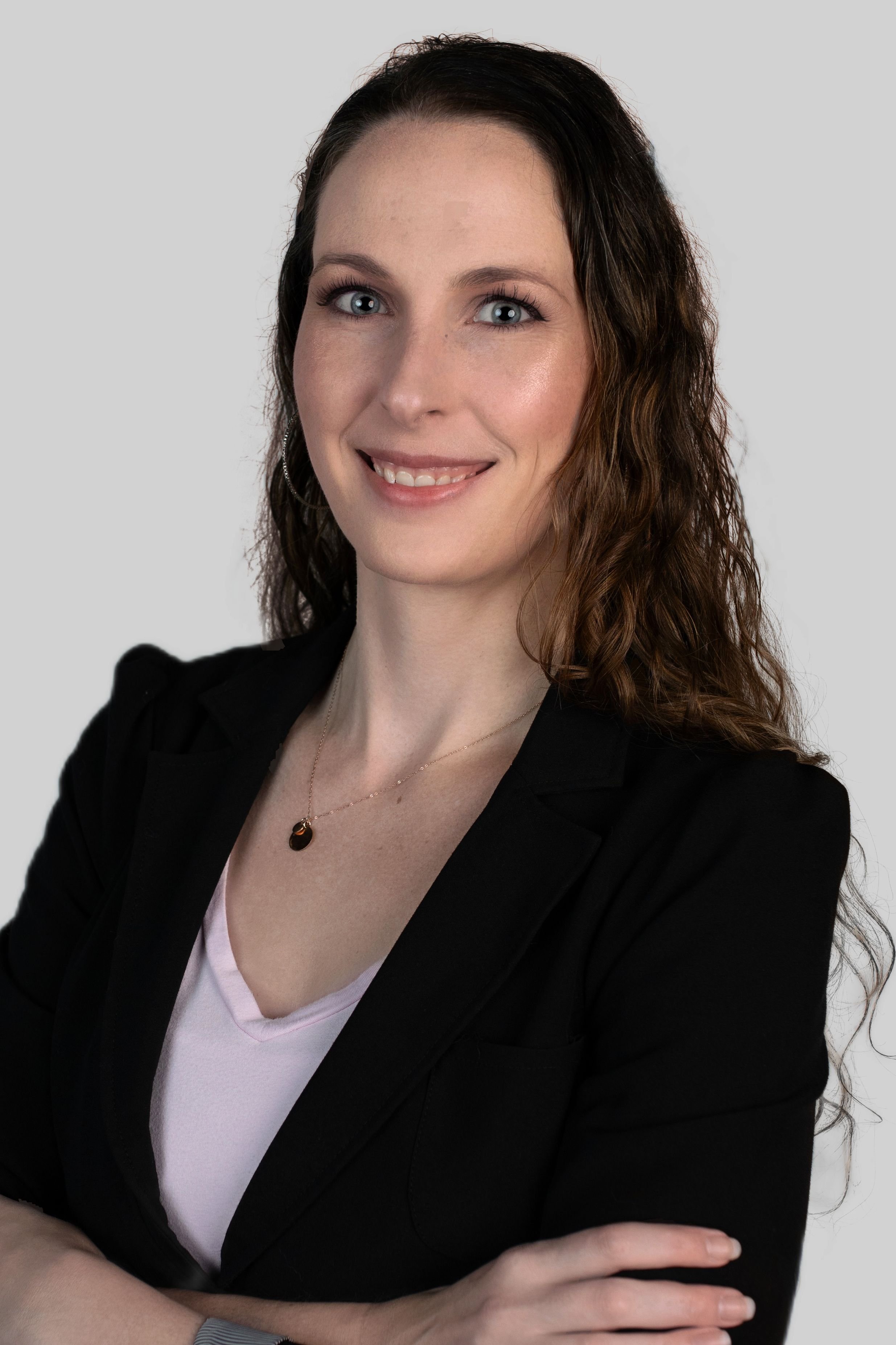A woman with wavy brown hair and blue eyes, wearing a black blazer over a pink top, smiling with her arms crossed against a plain light gray background.