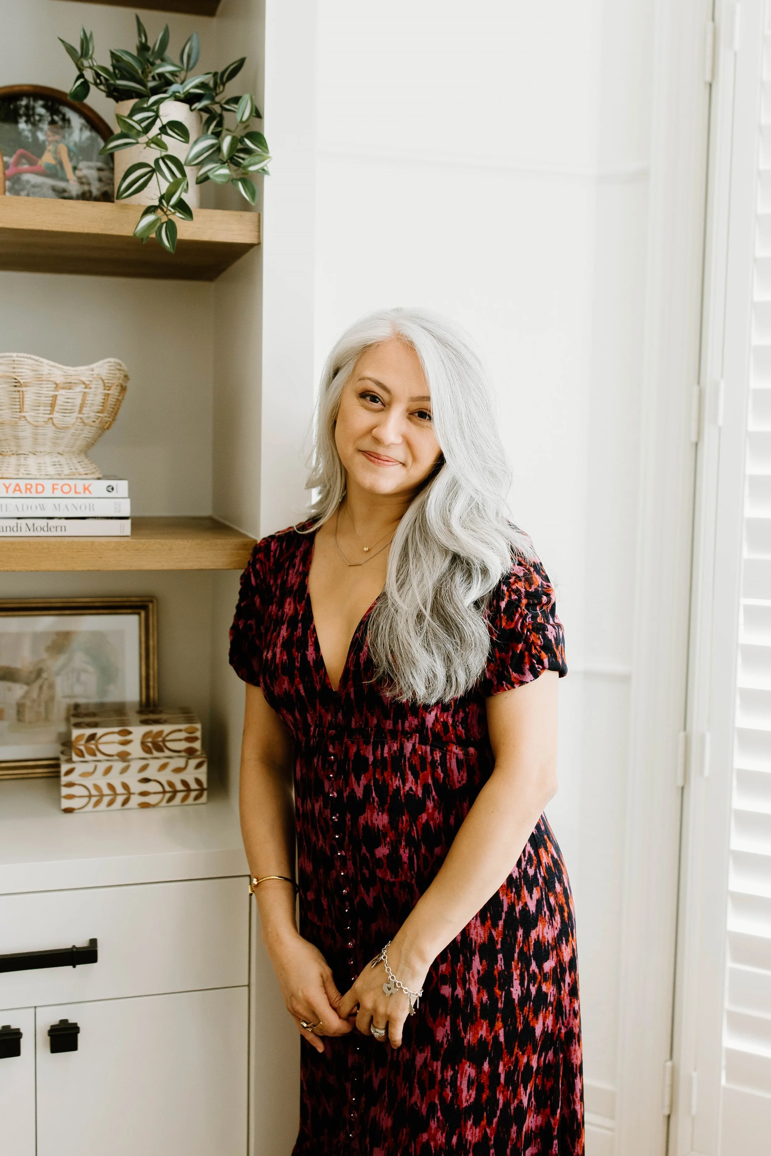 A woman with long, wavy gray hair smiling while standing indoors in front of a bookshelf with decorative items, using a red and black patterned dress.